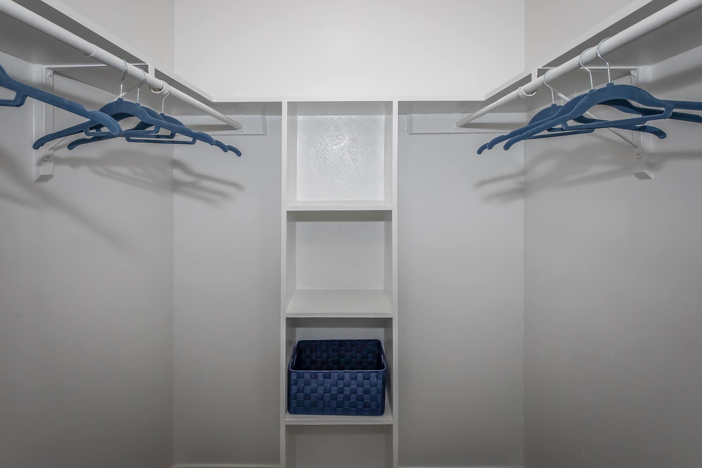 An empty closet with white walls and a light floor. Two clothing rods with blue hangers are mounted on each side. In the center, there is a small white shelving unit and a blue woven storage basket on the bottom shelf. The closet appears neat and organized, with a minimalistic design.