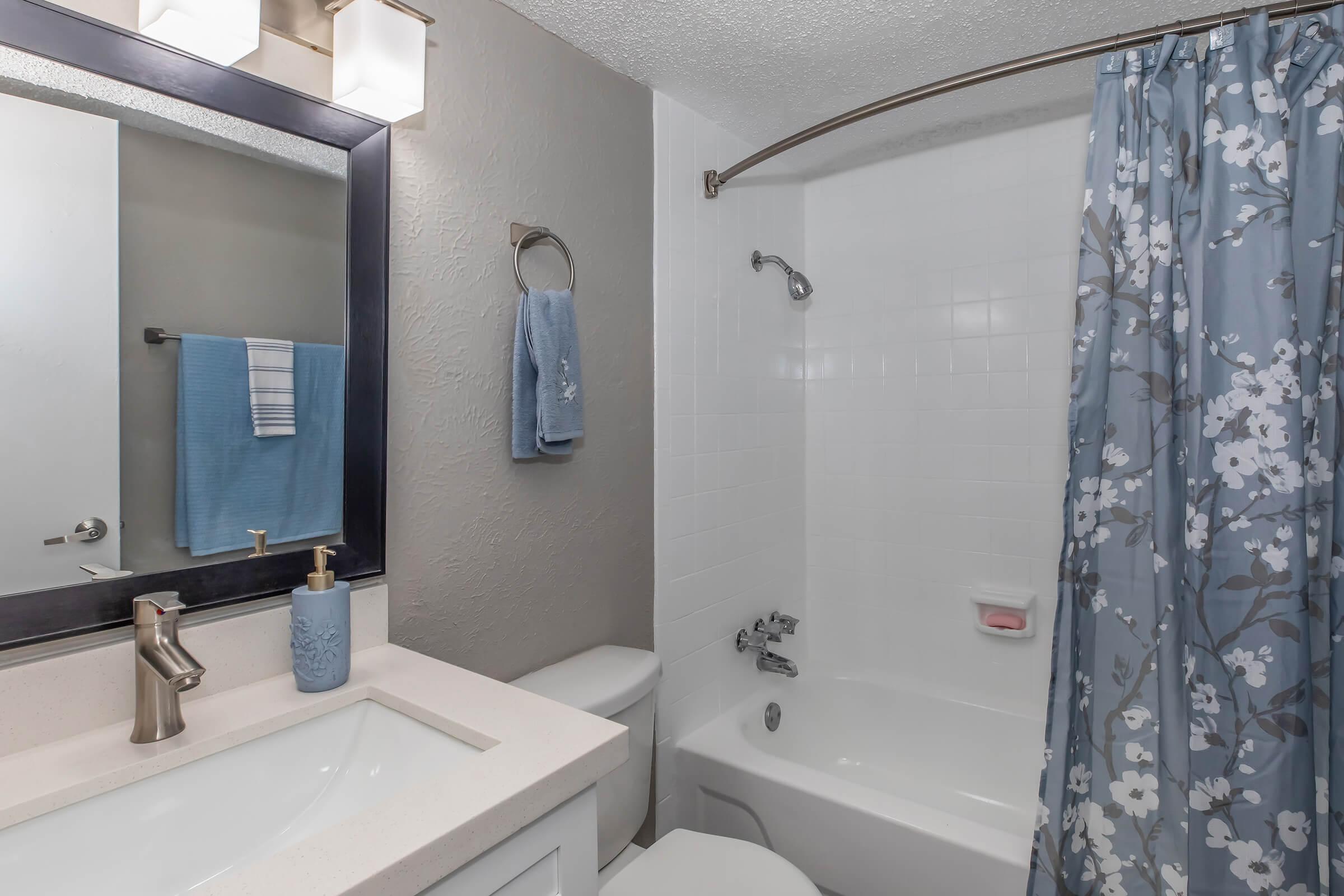 A modern bathroom featuring a shower/tub combo with a clear curtain adorned with a floral pattern. A white sink with a stylish faucet is visible, alongside a mirror and a towel rack holding a blue towel. The walls are painted in neutral tones, contributing to a clean and contemporary aesthetic.