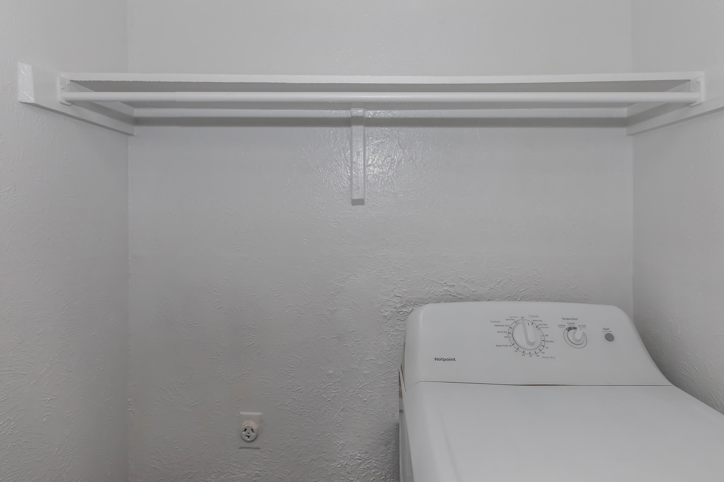 A clean, empty laundry room featuring a white washing machine on the right and a simple white shelving unit above it. The walls are painted in light gray, creating a bright and minimalist atmosphere, with no items on the shelves.