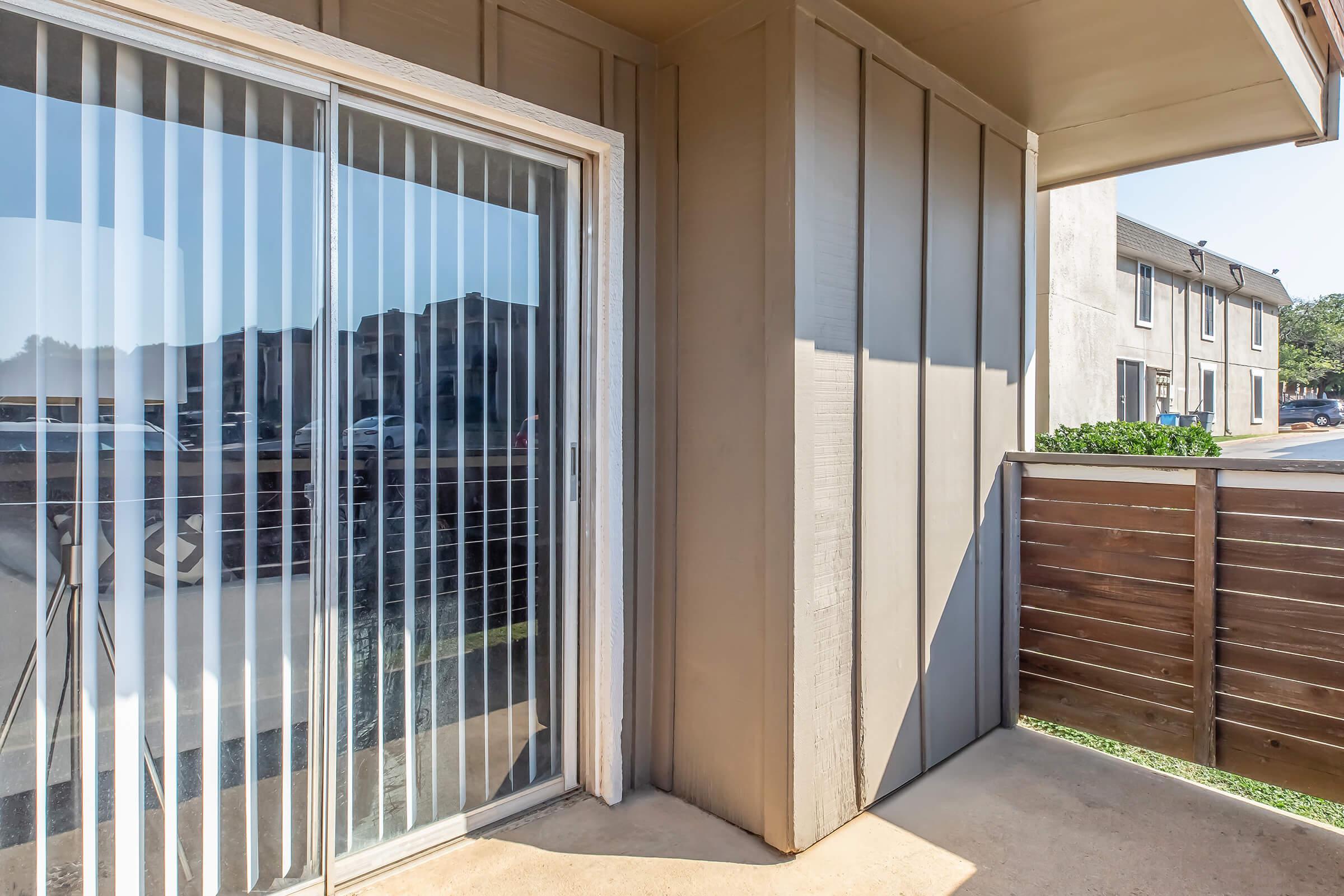 A corner view of a patio area featuring sliding glass doors with vertical blinds and a wooden fence. In the background, a two-story building is visible, along with parked cars. The setting appears to be part of an apartment community with bright daylight illuminating the space.