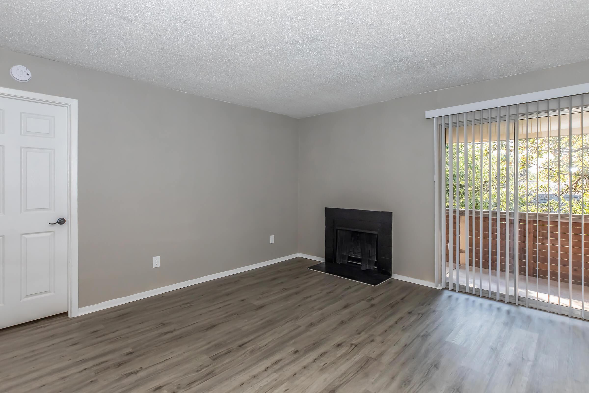 A spacious, neutral-colored living room with light gray walls and a wood-like floor. There is a black fireplace on one wall and a sliding glass door with vertical blinds that leads to a balcony, allowing natural light to fill the space.