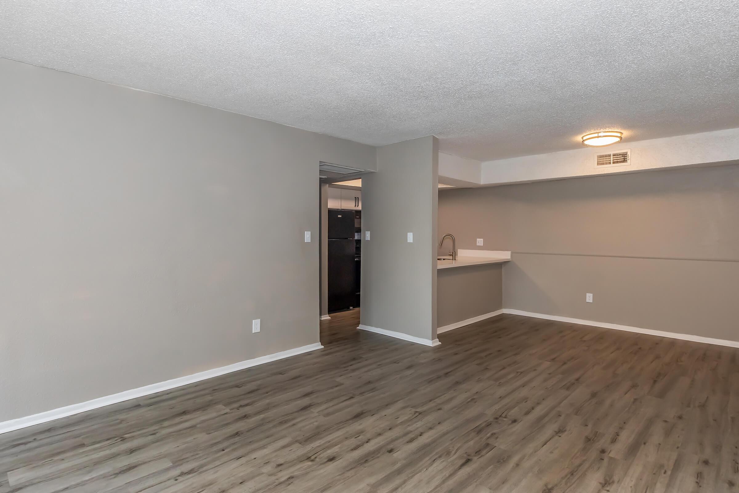 Spacious interior of a modern apartment with light gray walls and laminate flooring. A doorway leads to a kitchen area with a bar counter and a black refrigerator visible in the background. The room is well-lit, featuring a ceiling light fixture and an open floor plan, perfect for various furniture arrangements.