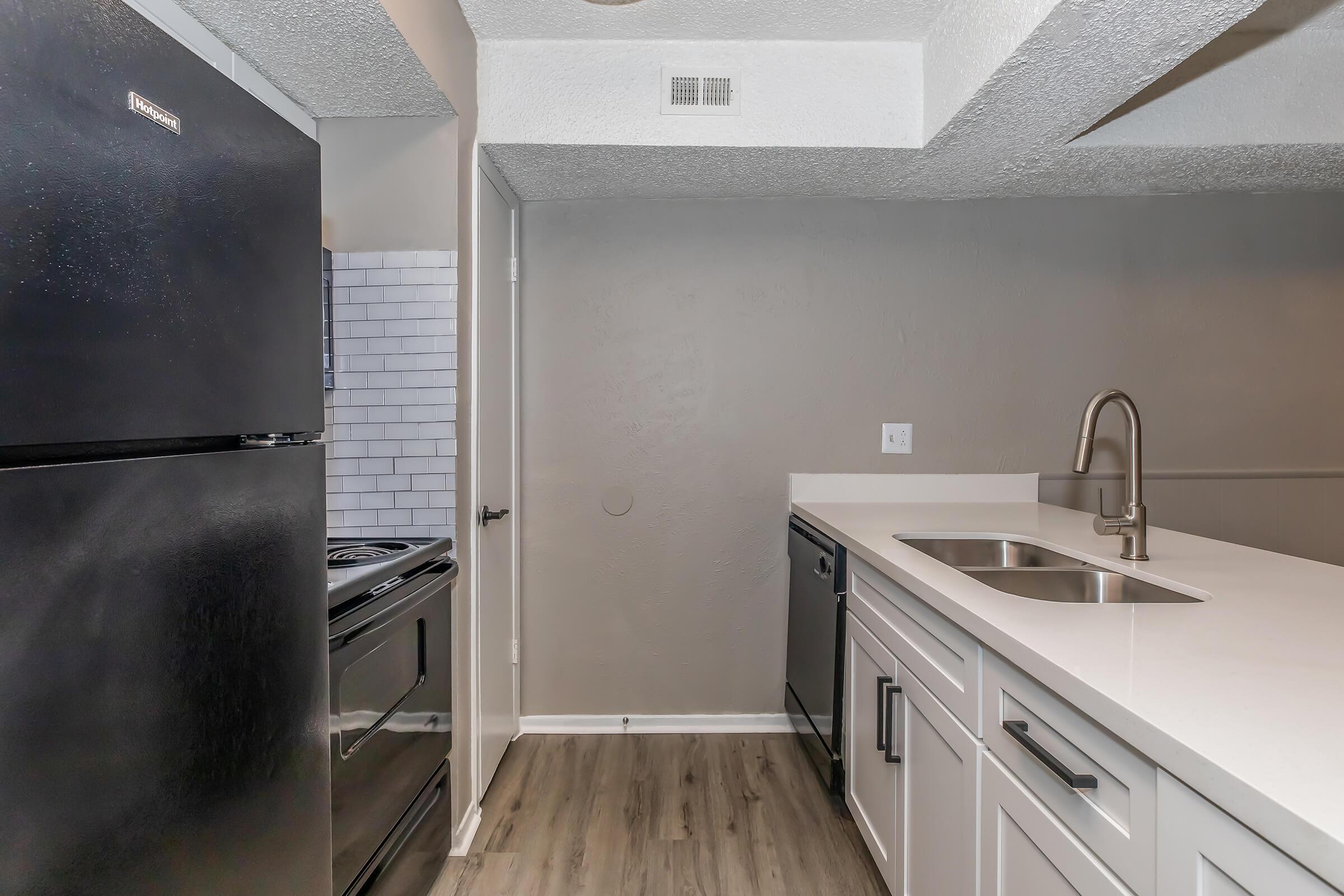 Modern kitchen featuring a black refrigerator and stove, white cabinets with a countertop, and a stainless steel sink. The walls are a neutral color, and there is a tile backsplash near the stove. The flooring is a light wood finish, providing a contemporary look.