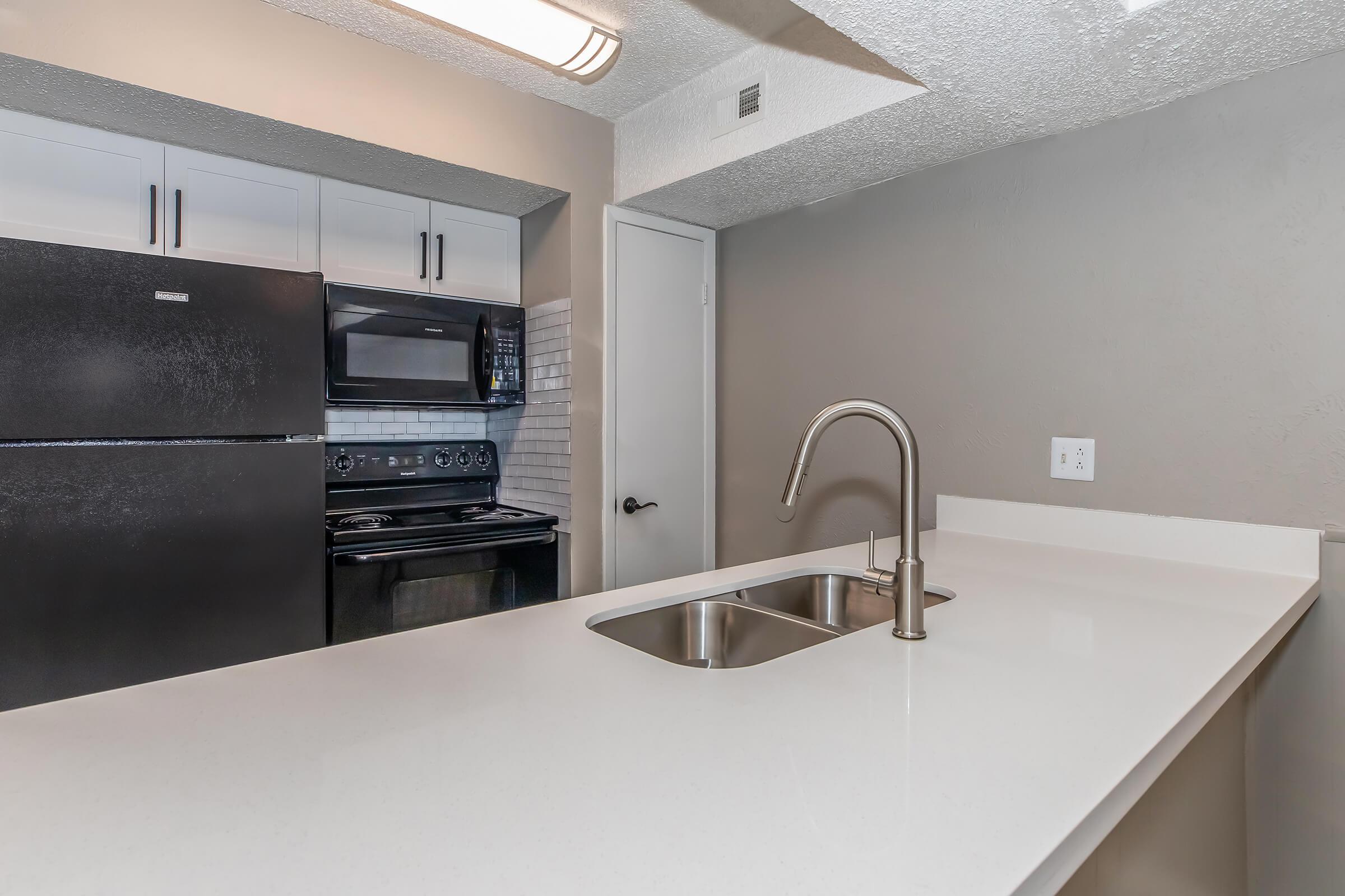 Modern kitchen featuring a black refrigerator, an oven with a stovetop, and a microwave. The countertops are white with a double sink. Cabinets above the counter are light-colored, and the walls are painted in a soft gray. A doorway leads to another room.