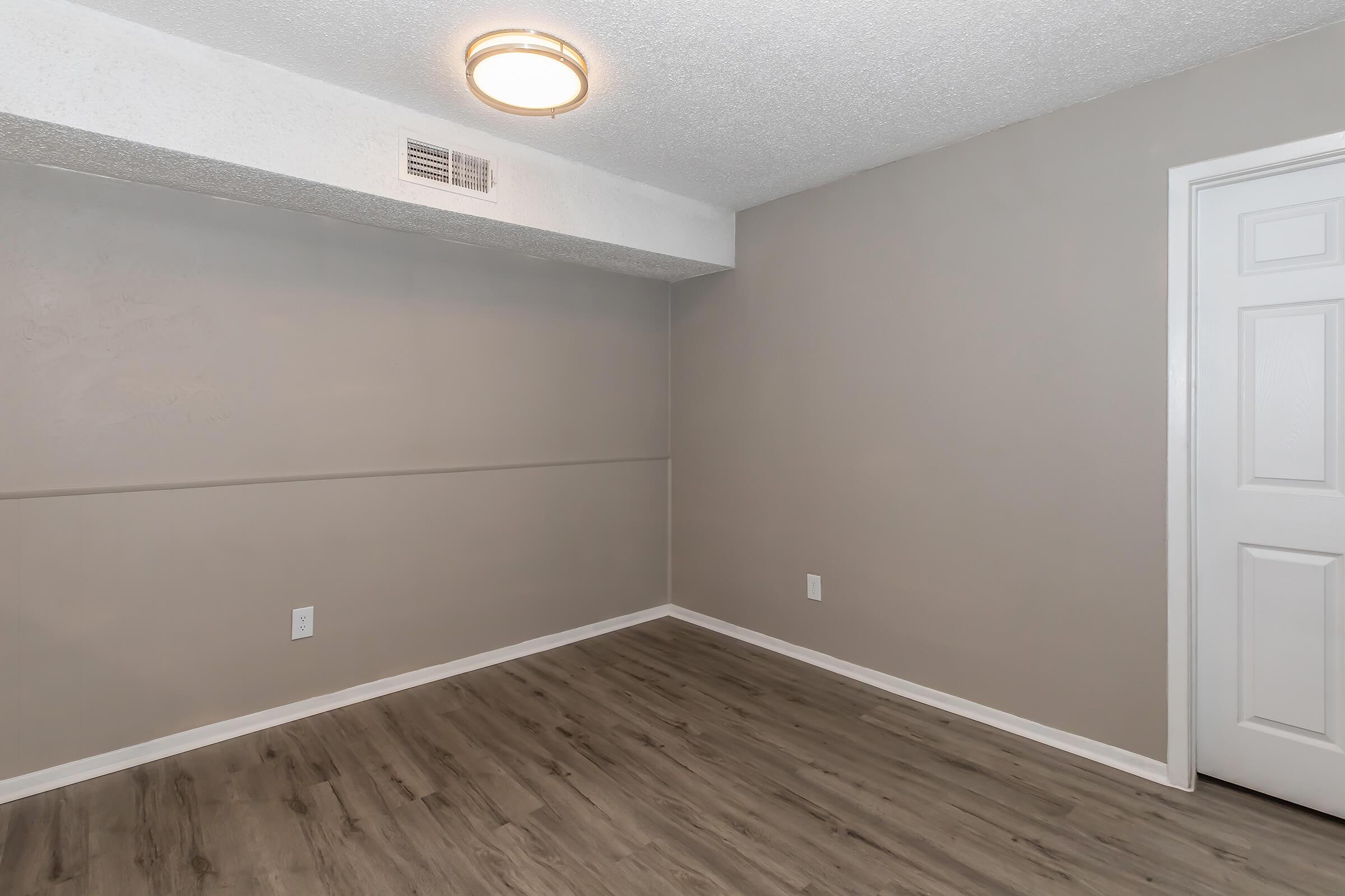 Empty room with light gray walls and wooden flooring. A simple ceiling light fixture illuminates the space. There is a white door on the right, and a small wall vent is visible above a horizontal line on the wall. The overall atmosphere is clean and modern.