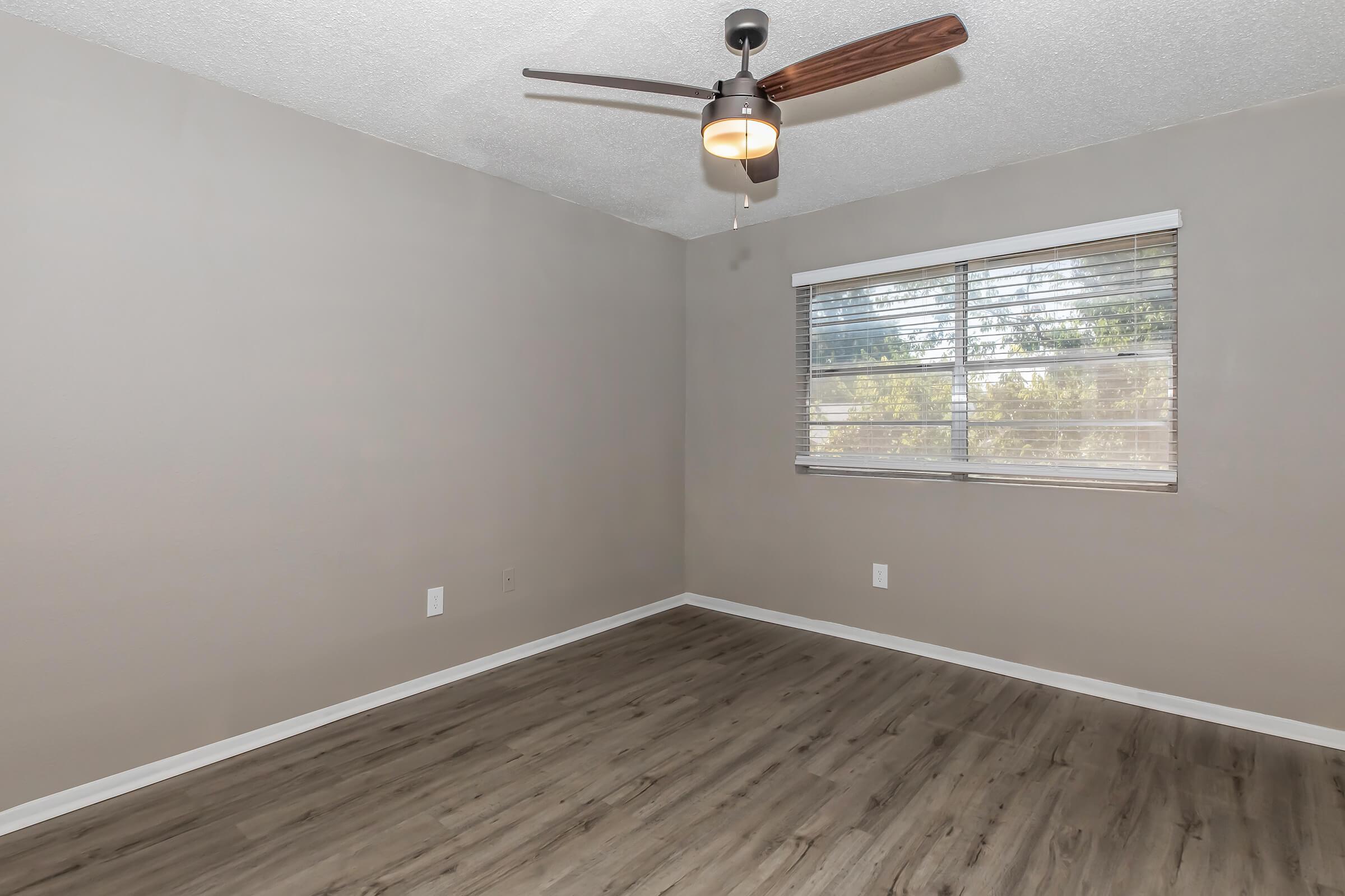 A well-lit, empty room featuring light gray walls, a ceiling fan with wooden blades, and a window with horizontal blinds. The flooring is a light-colored laminate, and there are no furnishings, creating a spacious, minimalist atmosphere.