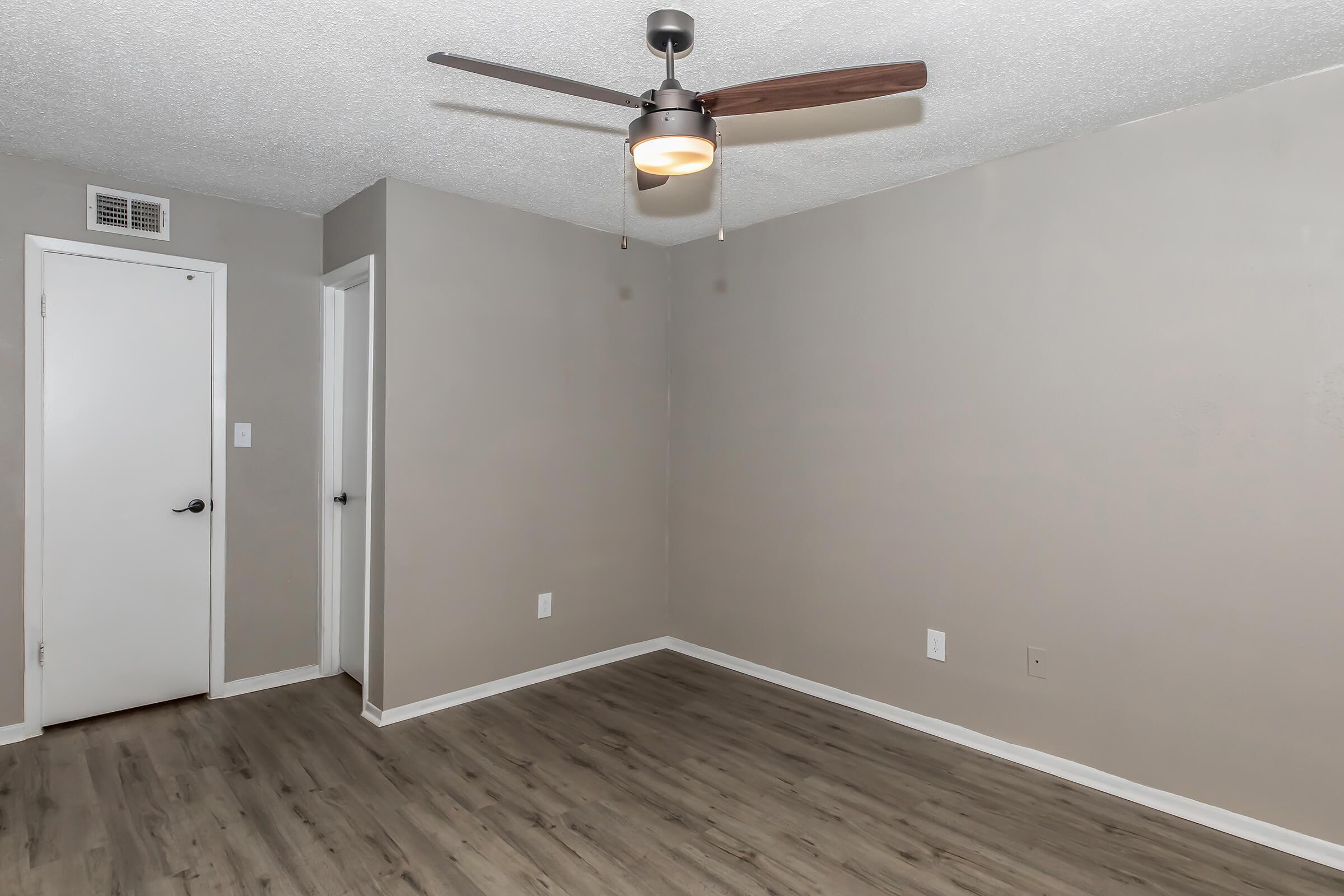 A spacious, empty room with light gray walls and a ceiling fan. The floor is wooden, and there are two doors visible—one to the left and one to the right. Natural light illuminates the area, creating a bright and airy atmosphere. Ideal for a living space or office setup.