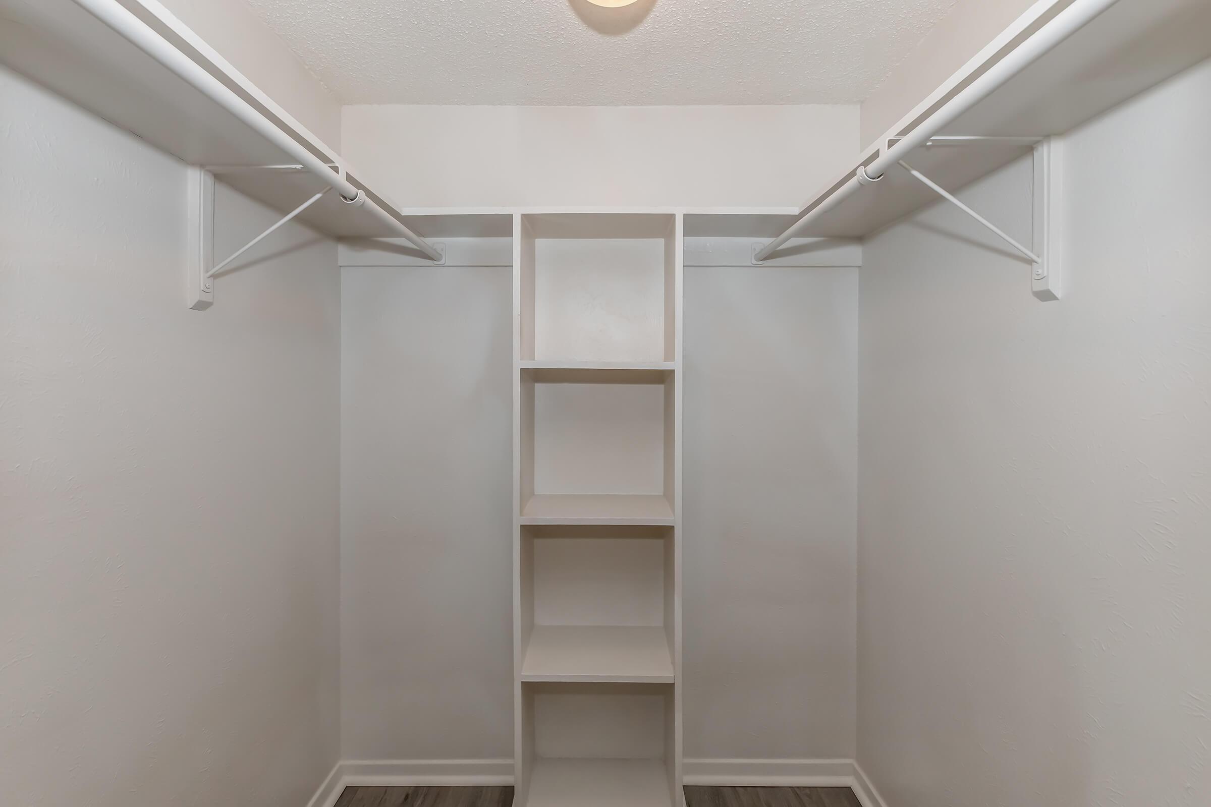 An empty walk-in closet featuring two hanging rods on either side and a central shelving unit with five open shelves. The walls are painted white, and the flooring is a light wood-style laminate. The ceiling has a light fixture. Overall, the space is uncluttered and bright.