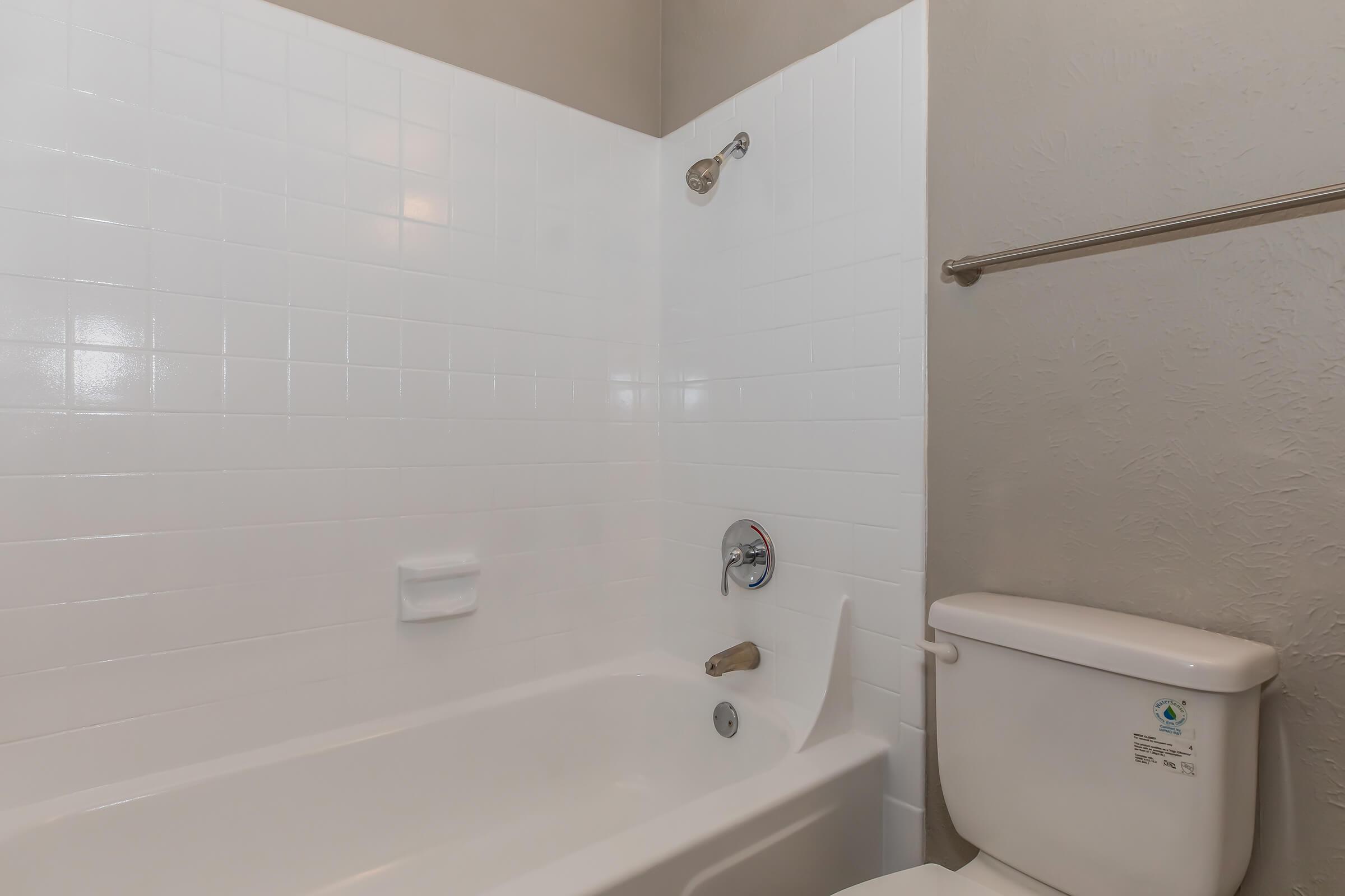 A clean, modern bathroom featuring a white tiled bathtub with a silver showerhead, a white toilet, and a towel rack against a beige wall. The space appears bright and well-maintained, highlighting the simplicity and functionality of the design.