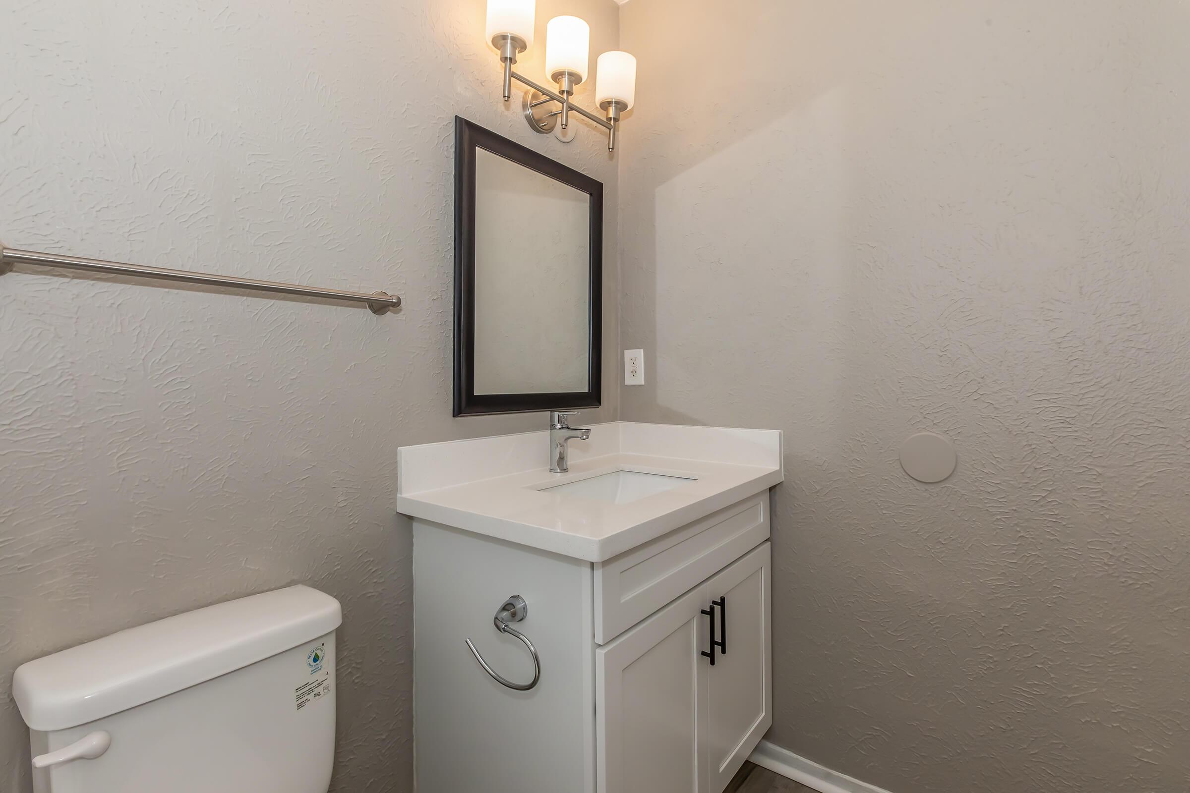 A small, modern bathroom featuring a white countertop sink with a black framed mirror above it. The walls are painted a light gray, and there are two light fixtures mounted above the mirror. A towel bar is attached to the wall, and a white toilet is located beside the sink. The flooring is simple and neutral.