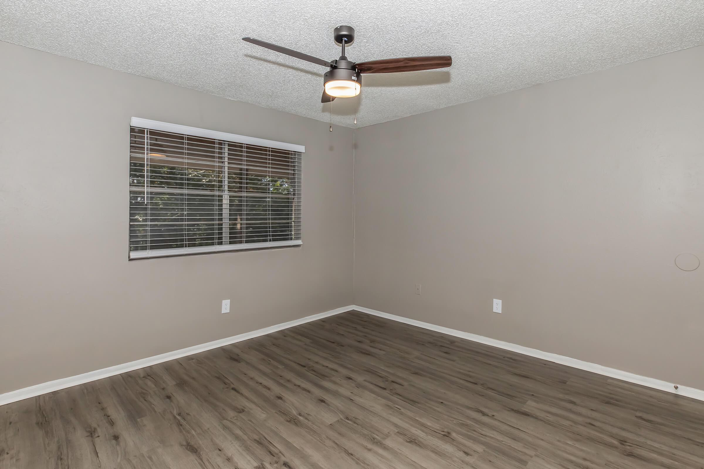 Empty room with light gray walls and a ceiling fan. A window with white blinds lets in natural light. Floor features light-colored wood laminate, creating a spacious and bright atmosphere. No furniture or decorations are present, emphasizing the room's size and simplicity.