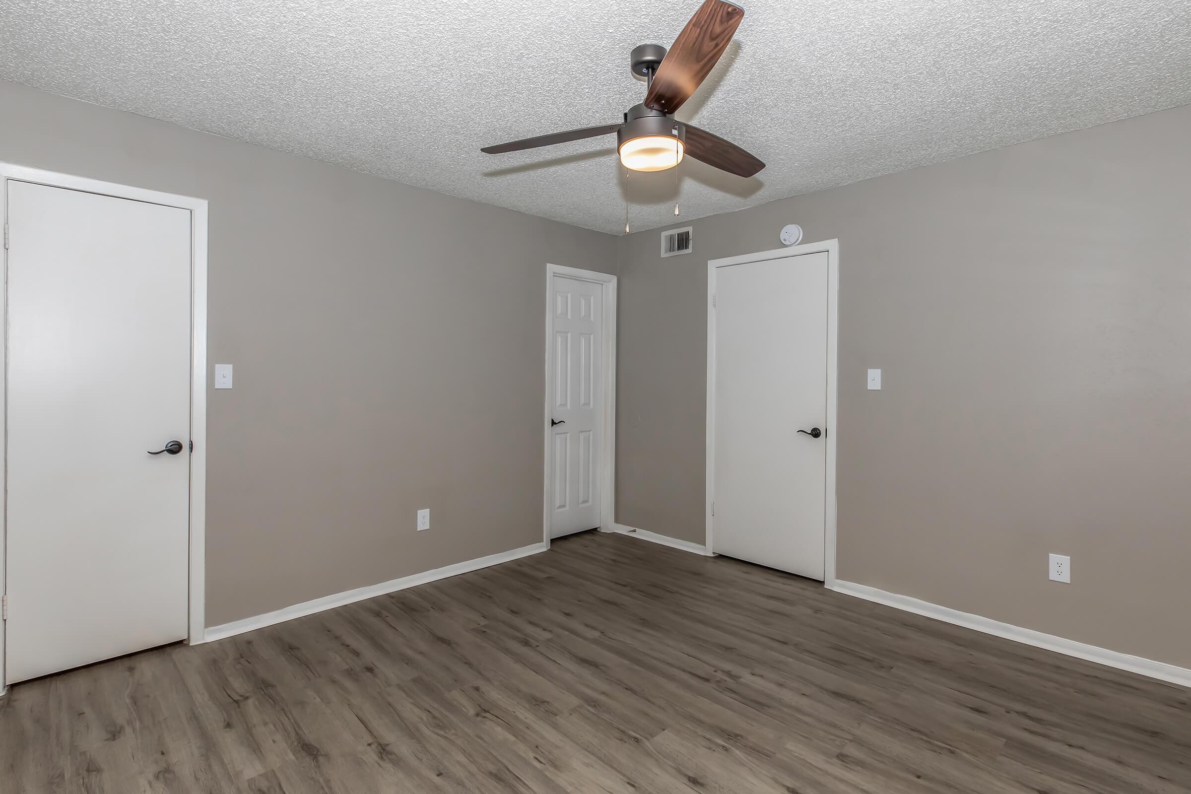 A spacious, empty room featuring light gray walls, a ceiling fan with a light fixture, and two closed white doors. The floor is a light wood laminate, contributing to a modern and airy feel. There are no visible furnishings or decorations, creating a clean and minimalist look.