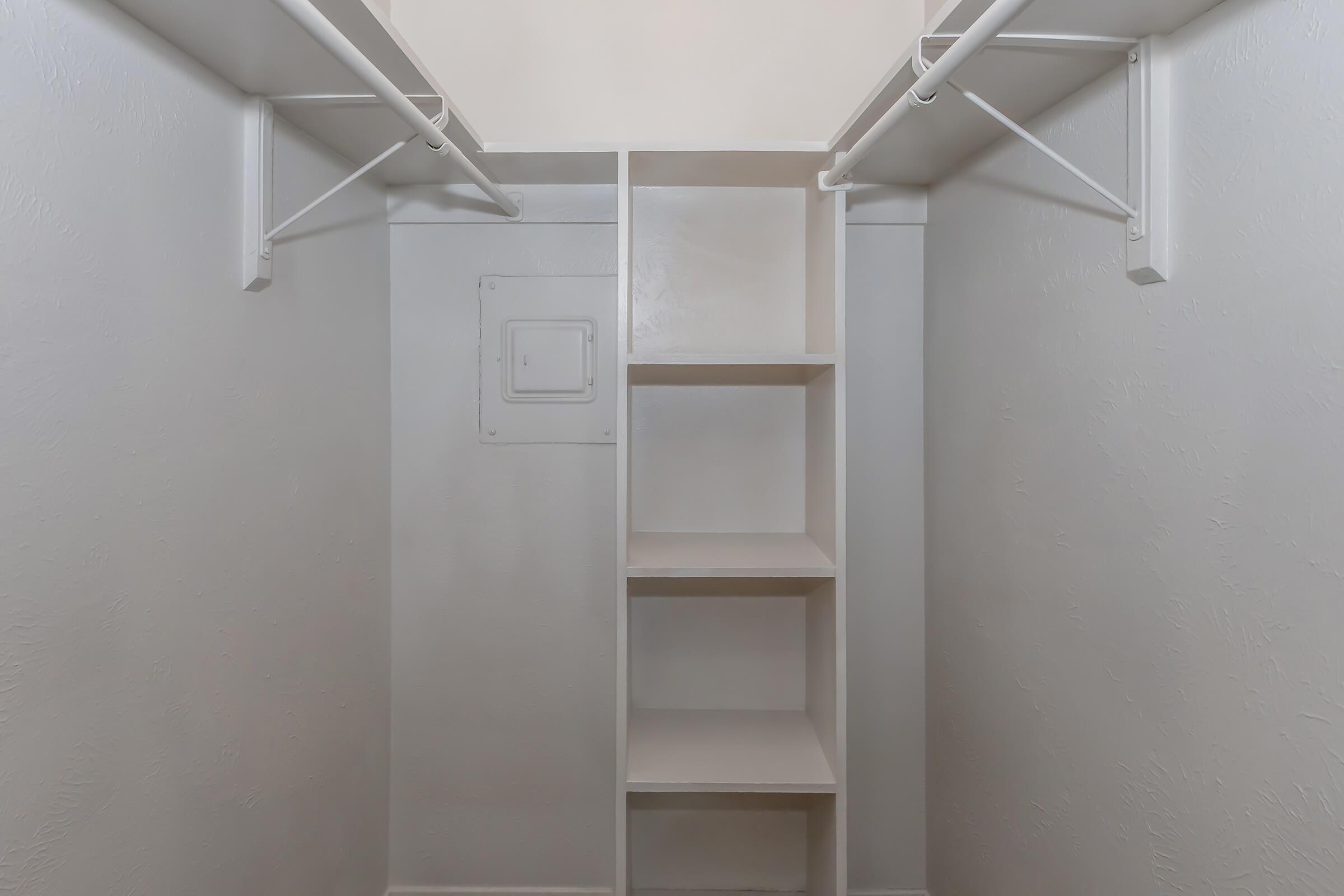 Empty closet with white walls and shelves. It features two hanging rods at the top and a vertical row of five open shelves on the right side. A small access panel is visible on the left wall. The overall appearance is clean and uncluttered.