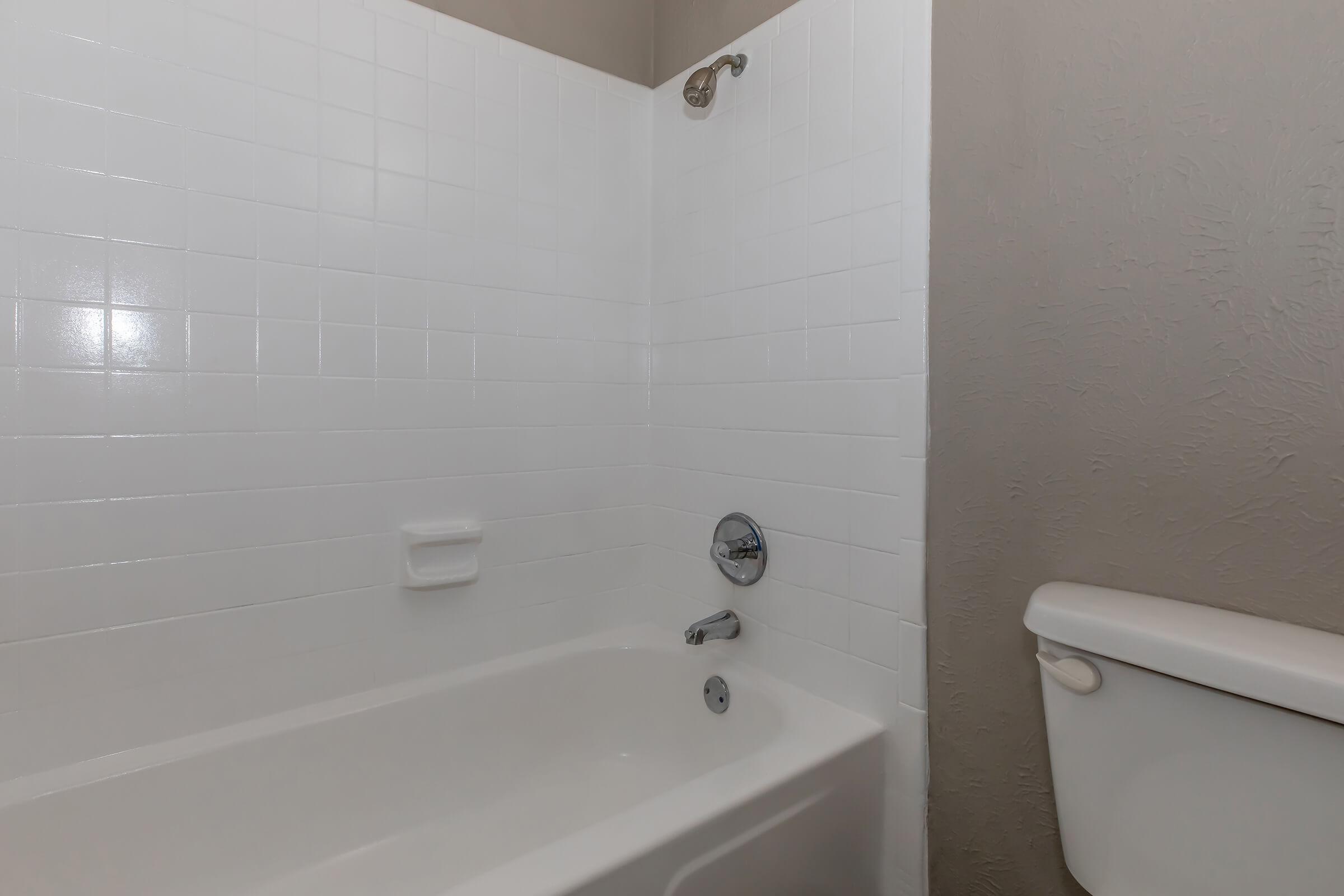 An interior bathroom view showcasing a clean, white tiled shower area with a showerhead, a bathtub, and a toilet. The walls are painted in a light neutral color, enhancing the bright and open atmosphere of the space.