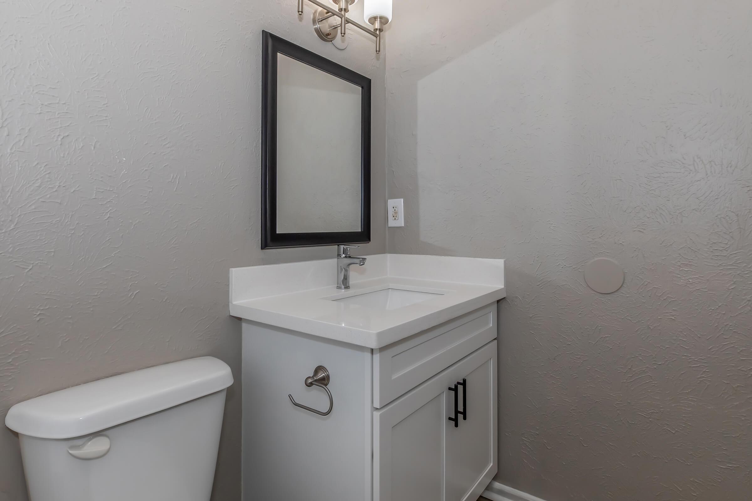 A modern bathroom featuring a white countertop sink with a mirror above, a wall-mounted towel holder, and a white toilet. The walls are painted in a light gray, and there is a light fixture above the mirror, creating a clean and contemporary look.