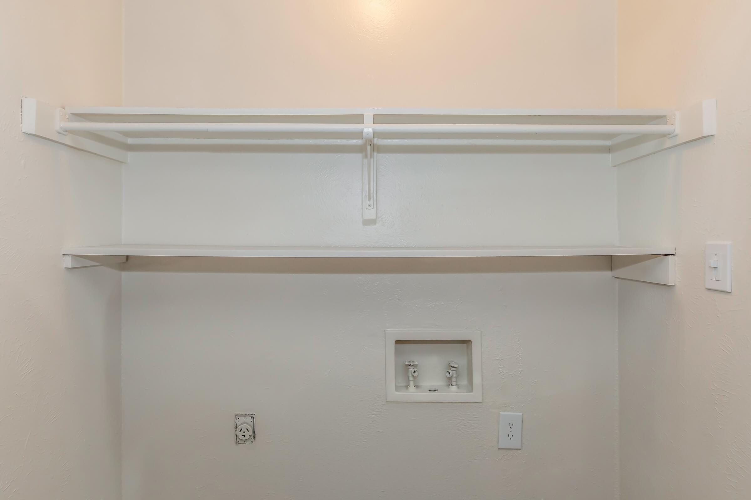 A clean, empty closet with two shelves and a white wall. The lower section features a small recessed area, likely for plumbing, with two fittings. There are light switches and an electrical outlet to the right. The overall design is simple and modern, with neutral colors.