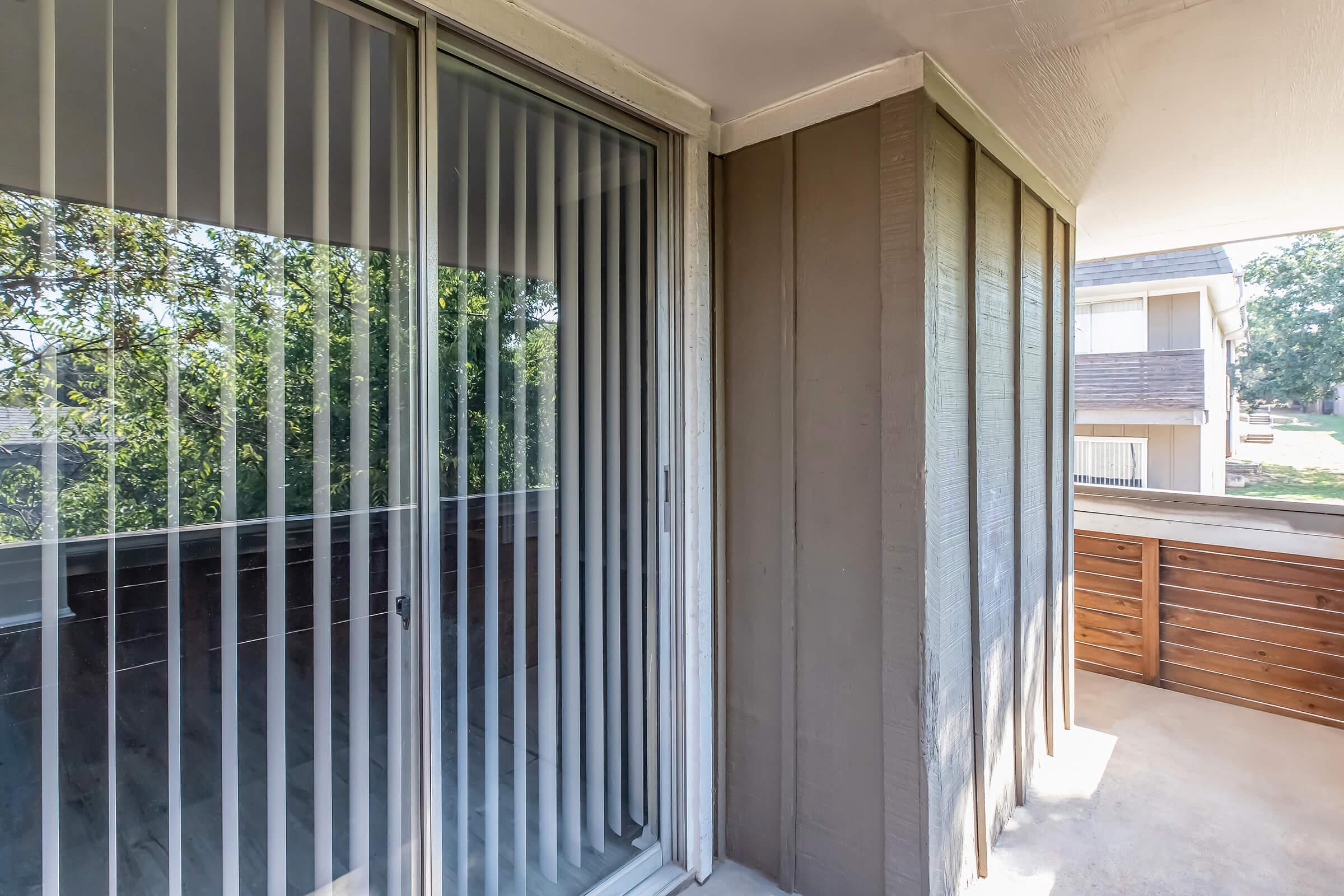 A modern balcony featuring vertical blinds on glass sliding doors. The exterior walls are a combination of light and dark wood paneling. In the background, there are glimpses of greenery and another building, indicating a residential setting. The floor is concrete, providing a clean, minimalist look.