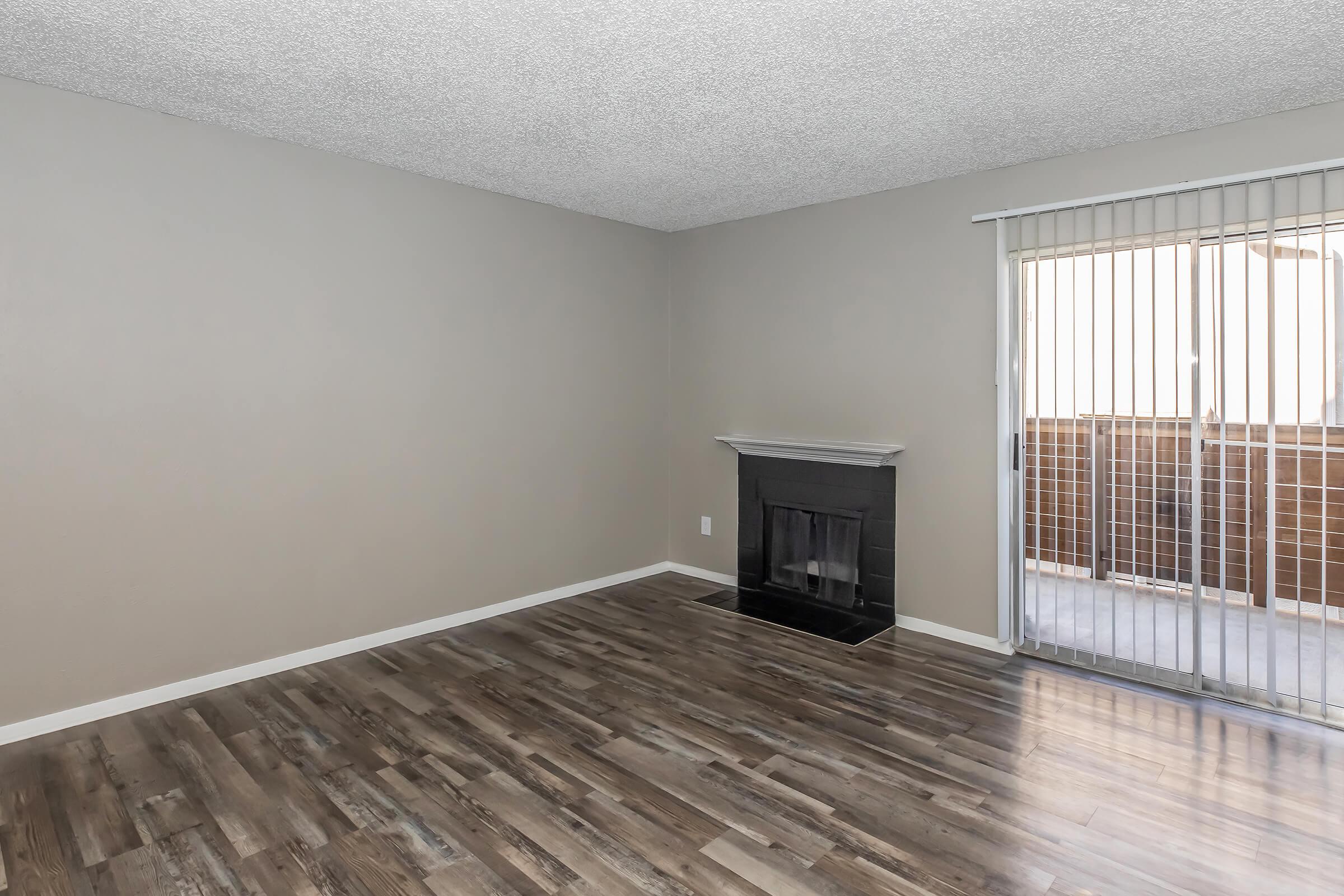 A spacious, empty living room with light gray walls and a textured ceiling. The room features a dark fireplace on one side and sliding glass doors leading to a patio. The floor is finished with wide, light-colored wooden planks, creating an inviting atmosphere.