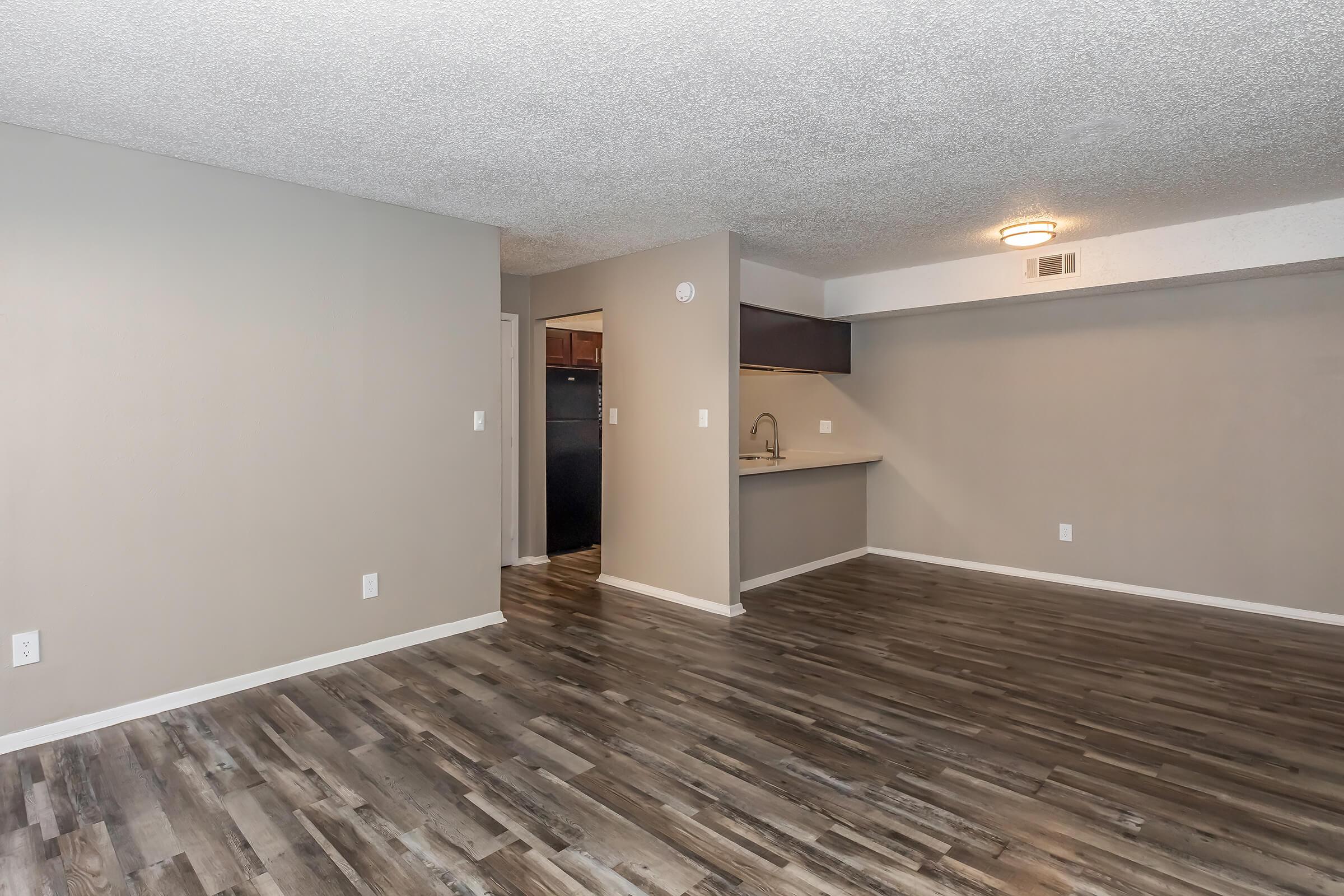 A spacious, empty living area featuring laminate flooring, textured ceiling, and neutral-colored walls. A kitchen is partially visible in the background, showcasing a bar counter with a sink. The room is well-lit with a ceiling light fixture.