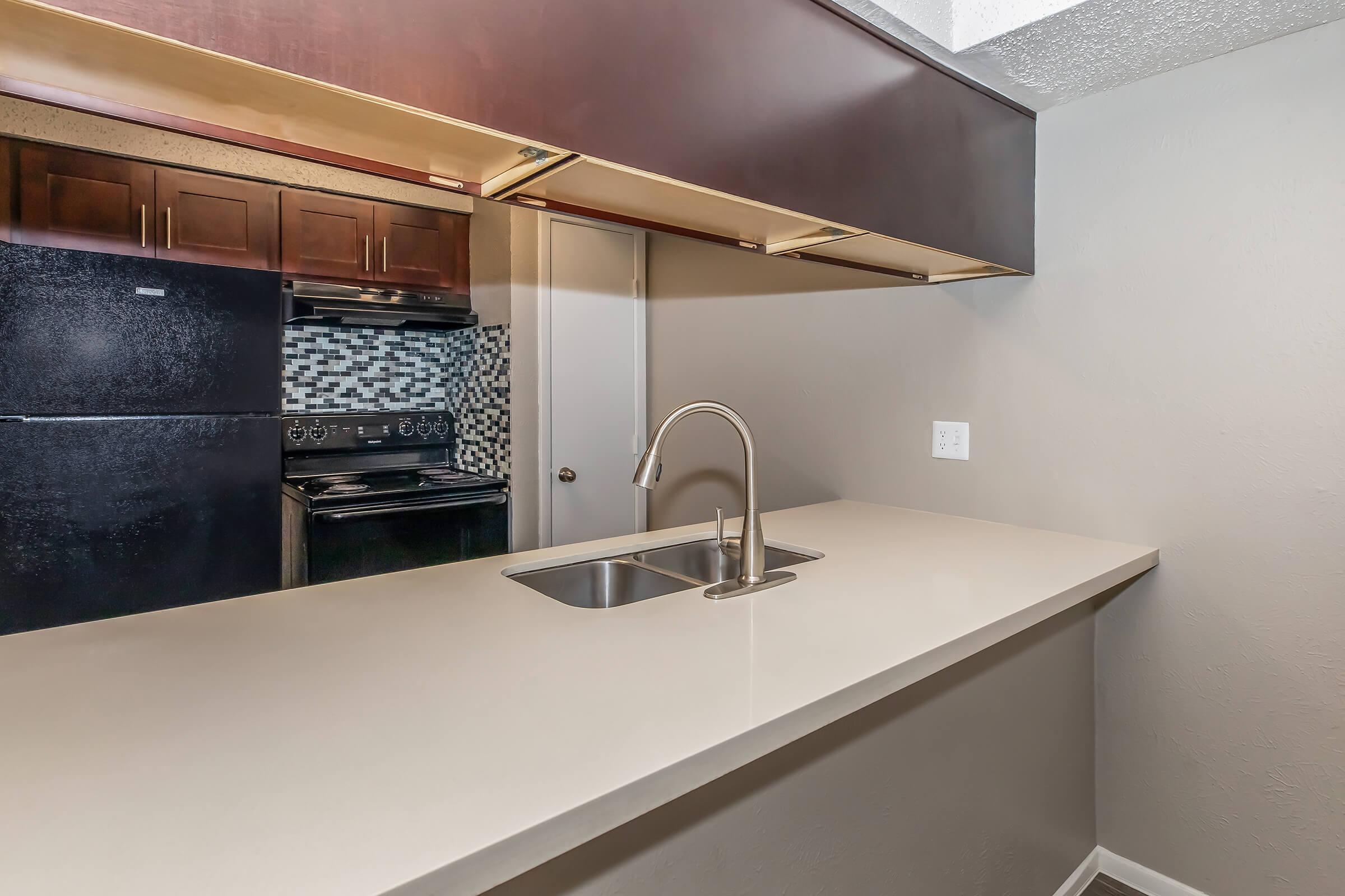 Modern kitchen featuring a sleek countertop with a single sink, dark wooden cabinets, and stainless steel appliances. A black refrigerator and an oven are visible, along with a stylish backsplash. The space is well-lit with neutral wall colors, creating a contemporary and inviting atmosphere.