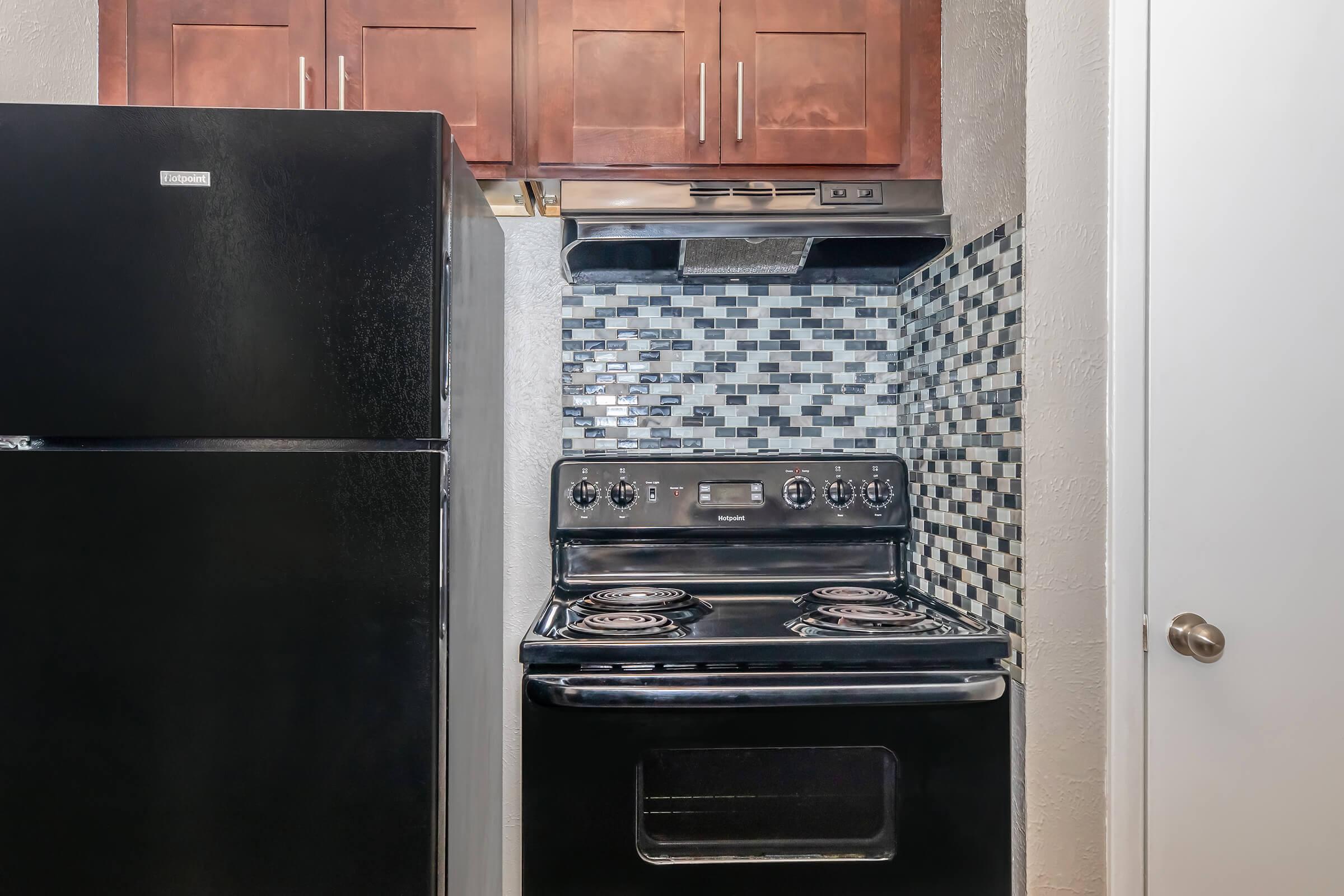 A modern kitchen featuring a black refrigerator next to a black stove with an oven. The stove is equipped with four burners and an overhead range hood. The backsplash is made of small, decorative tiles in shades of black, gray, and white, complemented by wooden cabinets above.