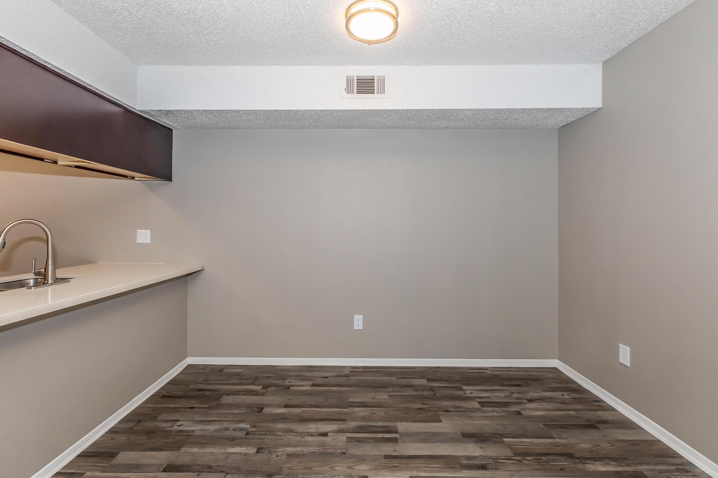 A vacant room with gray walls and a textured ceiling. It features a small kitchen area with a countertop and dark cabinetry to the left. The flooring is wood-like, and there is a round ceiling light. The space appears to be well-lit, ready for furnishing.