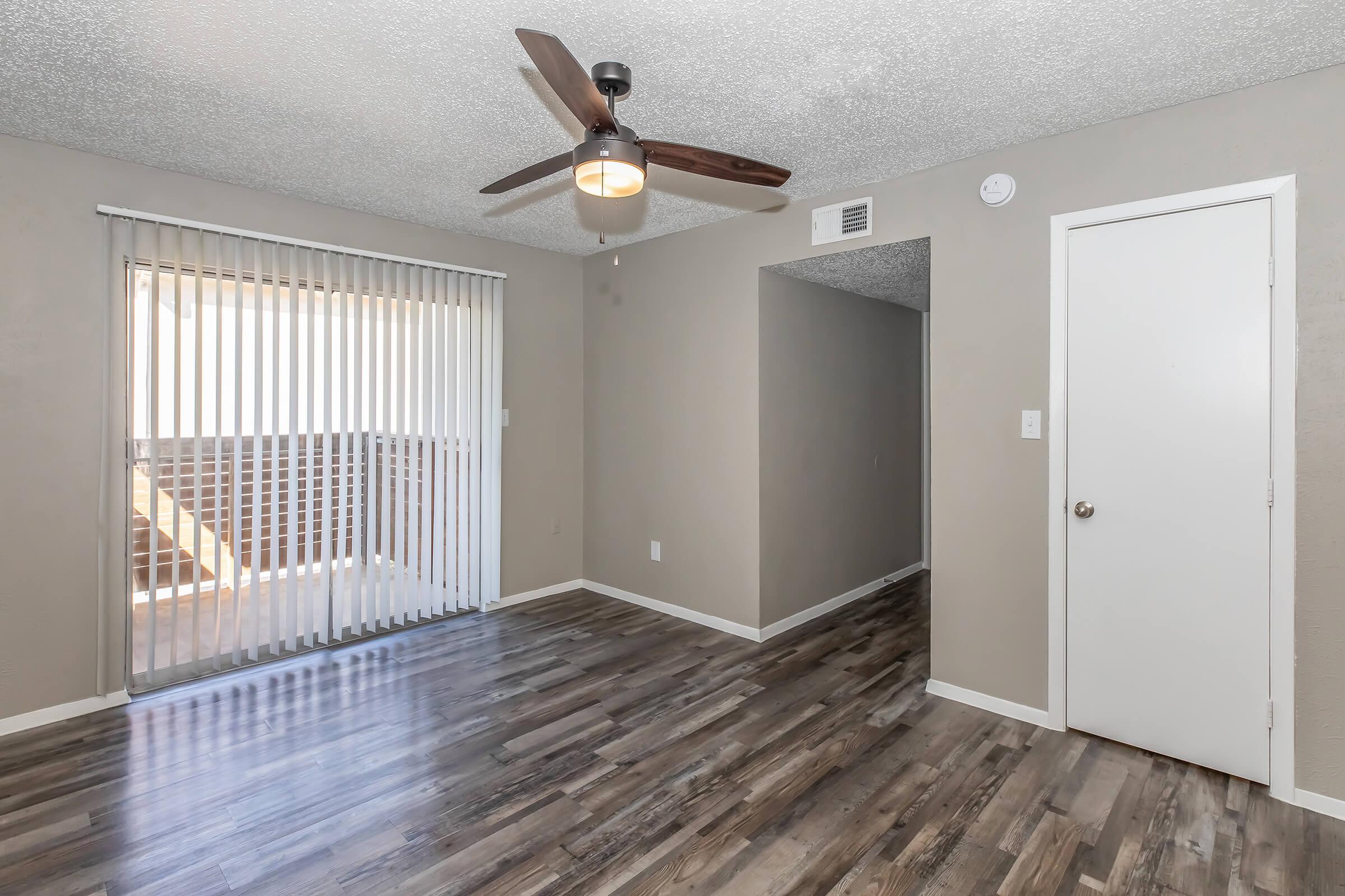 A spacious, well-lit room featuring a ceiling fan, laminate flooring, and sliding glass doors with vertical blinds. A doorway leads to another area, and the walls are painted a neutral color, enhancing the open feel of the space.