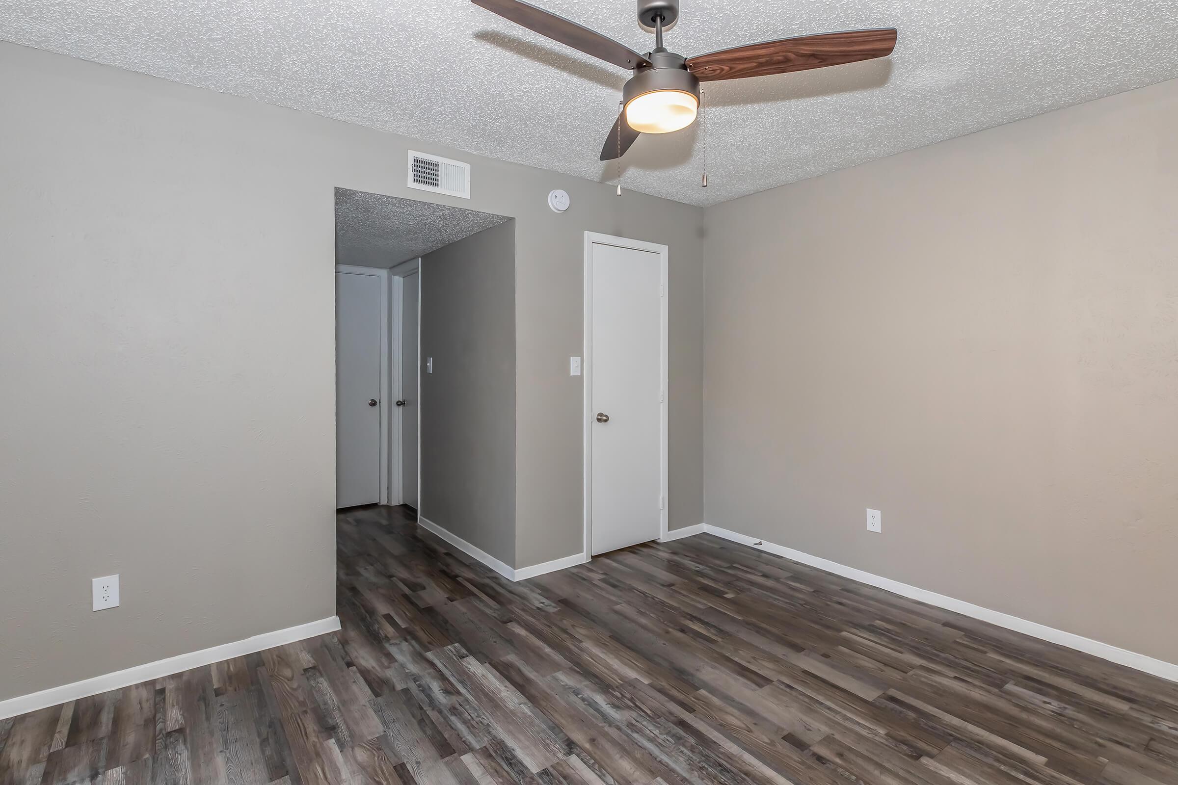 A well-lit, empty room featuring textured gray walls and a ceiling fan with wooden blades. The floor is covered in dark wood laminate. A doorway leads to another room on the left, and a closed door is visible on the right. The space appears clean and ready for furnishings.