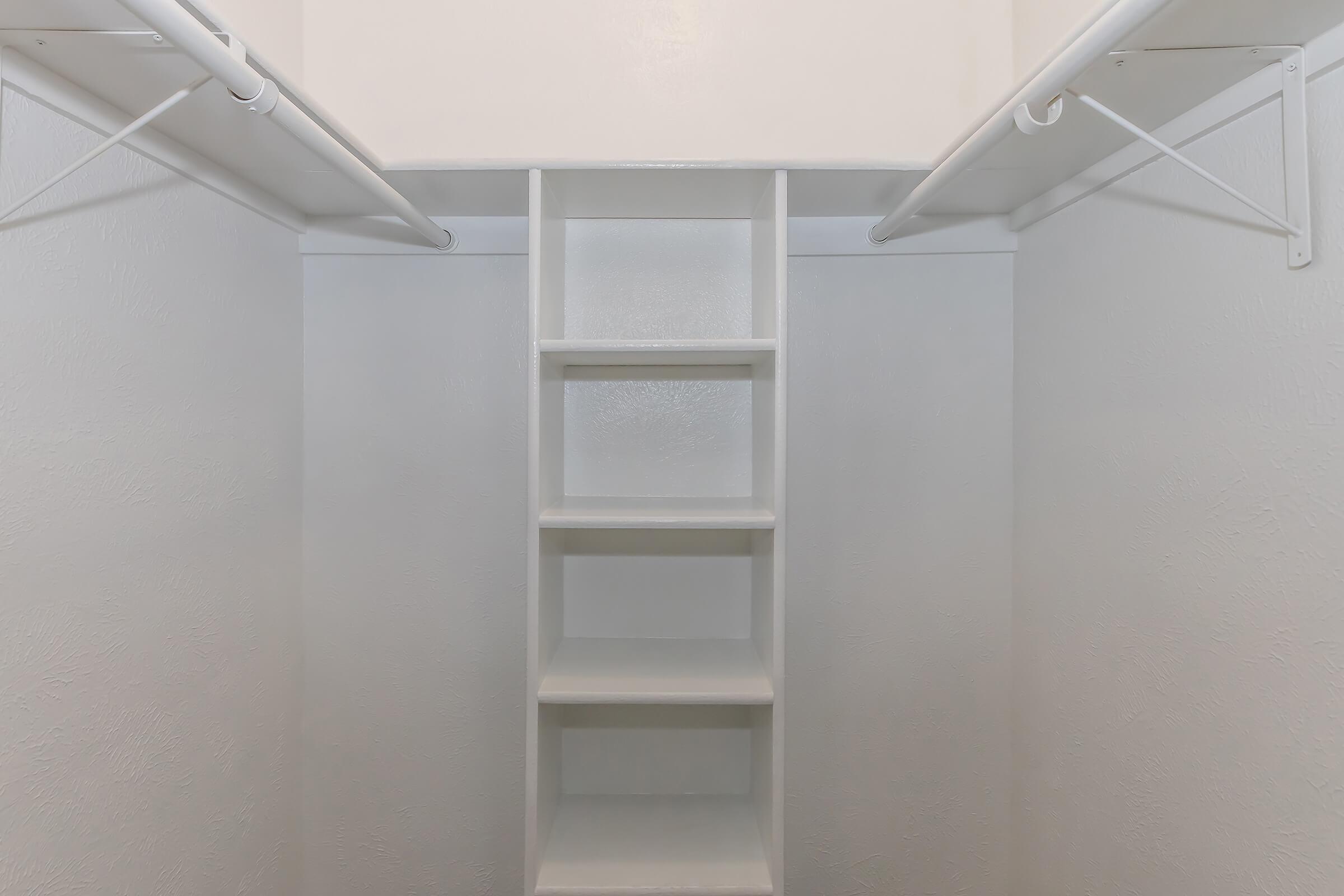 Empty white closet with two hanging rods at the top and a set of five shelves along the back. A minimalist design featuring clean lines and a bright, neutral color scheme, emphasizing the spaciousness of the area.