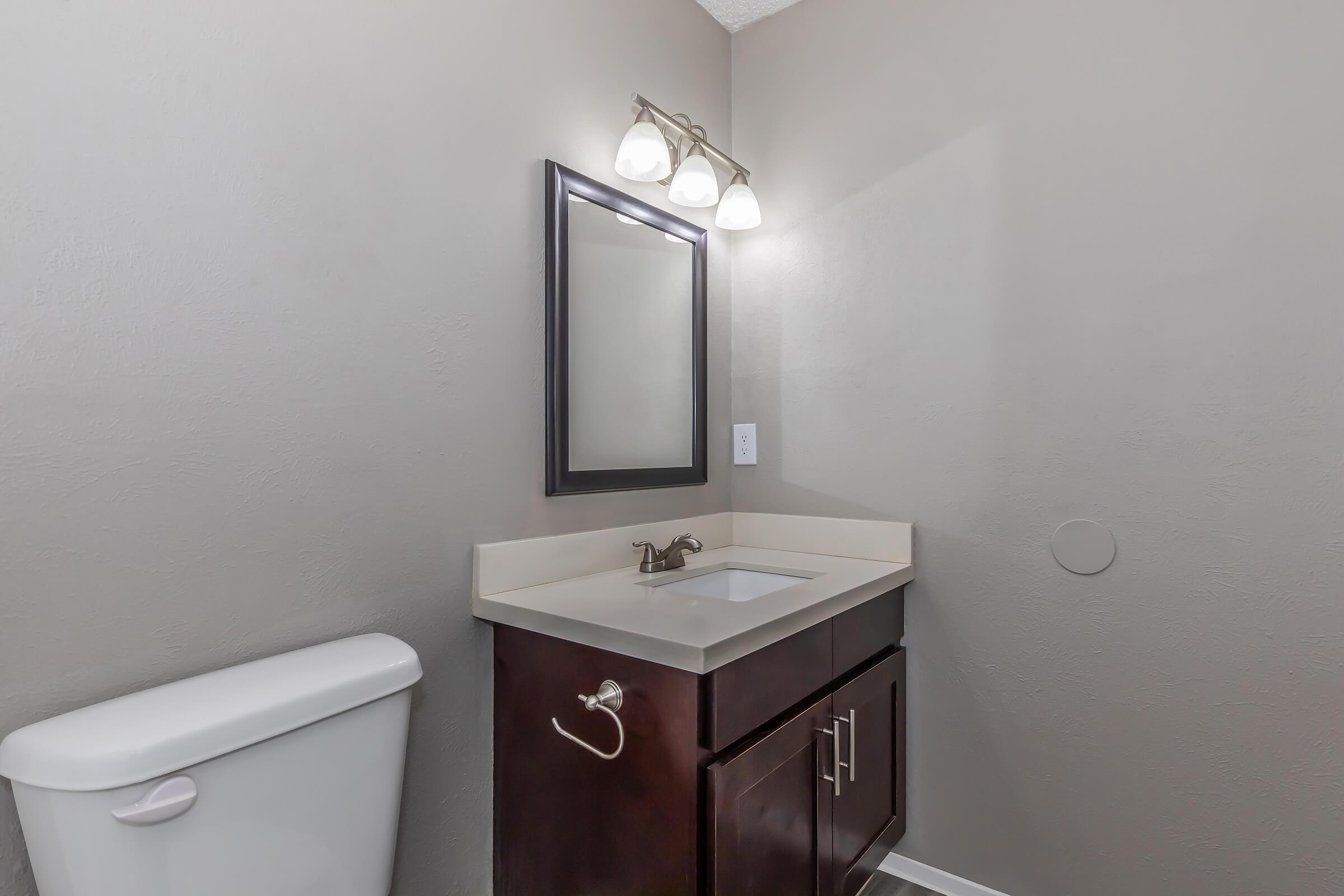 A clean and modern bathroom featuring a white toilet, a dark wood vanity with a sink, and a large mirror above it. The walls are painted in a neutral gray color, and there are three light fixtures mounted above the mirror, providing bright illumination. The overall aesthetic is simple and functional.