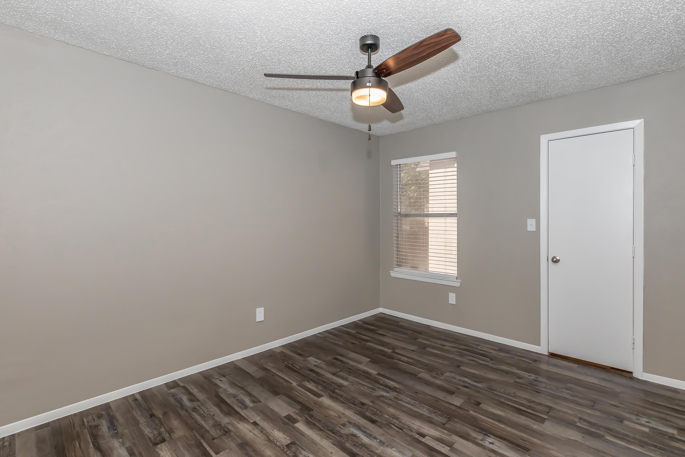 A spacious, empty room featuring a light-colored wall, a ceiling fan with wooden blades, and a large window allowing natural light. The floor is covered in wooden laminate, and there is a closed white door on one wall, enhancing the modern and minimalistic aesthetic of the space.