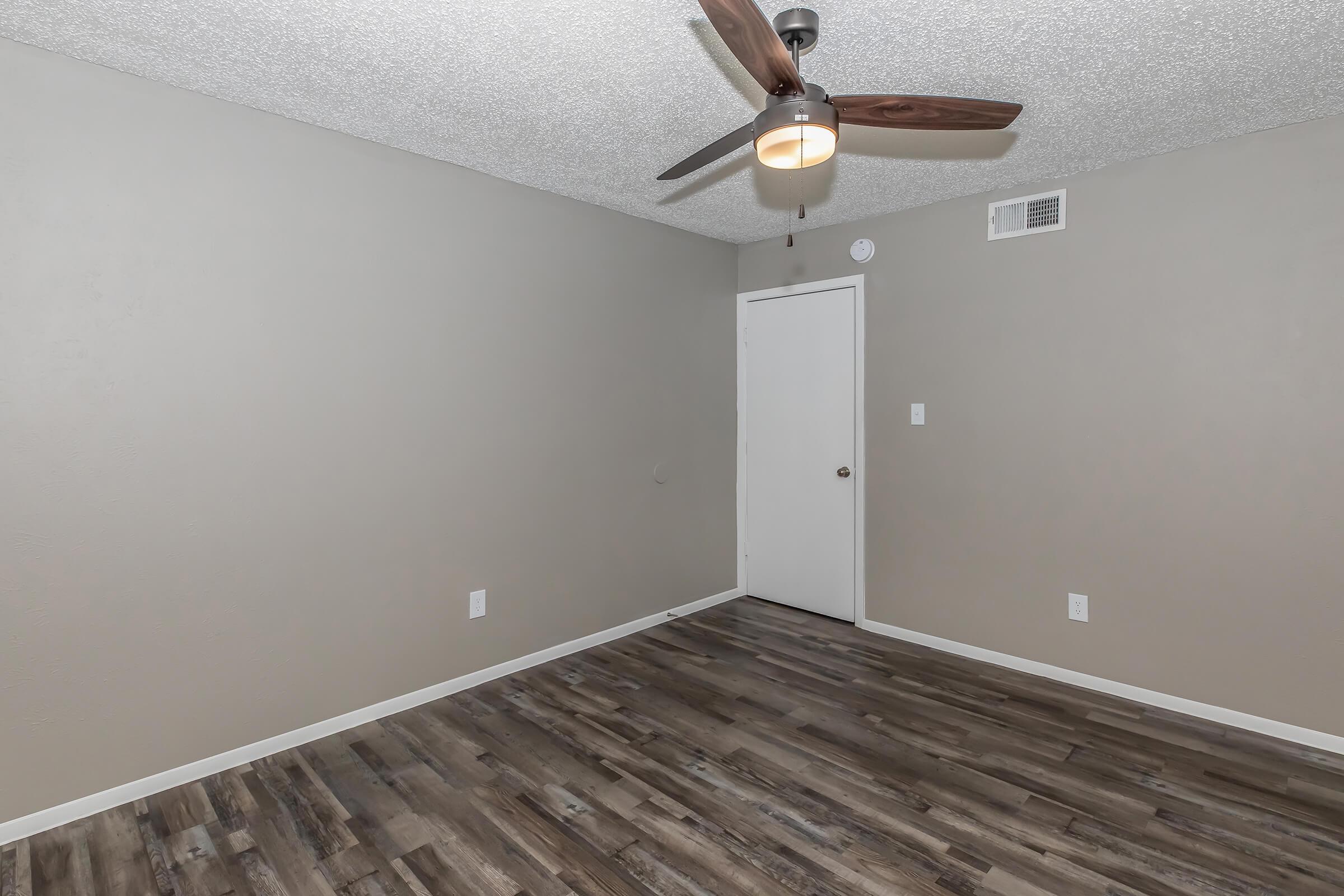 An empty room with light gray walls and a ceiling fan. The flooring is wood-like laminate in shades of brown. A closed white door is visible on one wall, with electrical outlets and a vent located nearby. The space is bright and uncluttered, giving a modern and minimalist appearance.