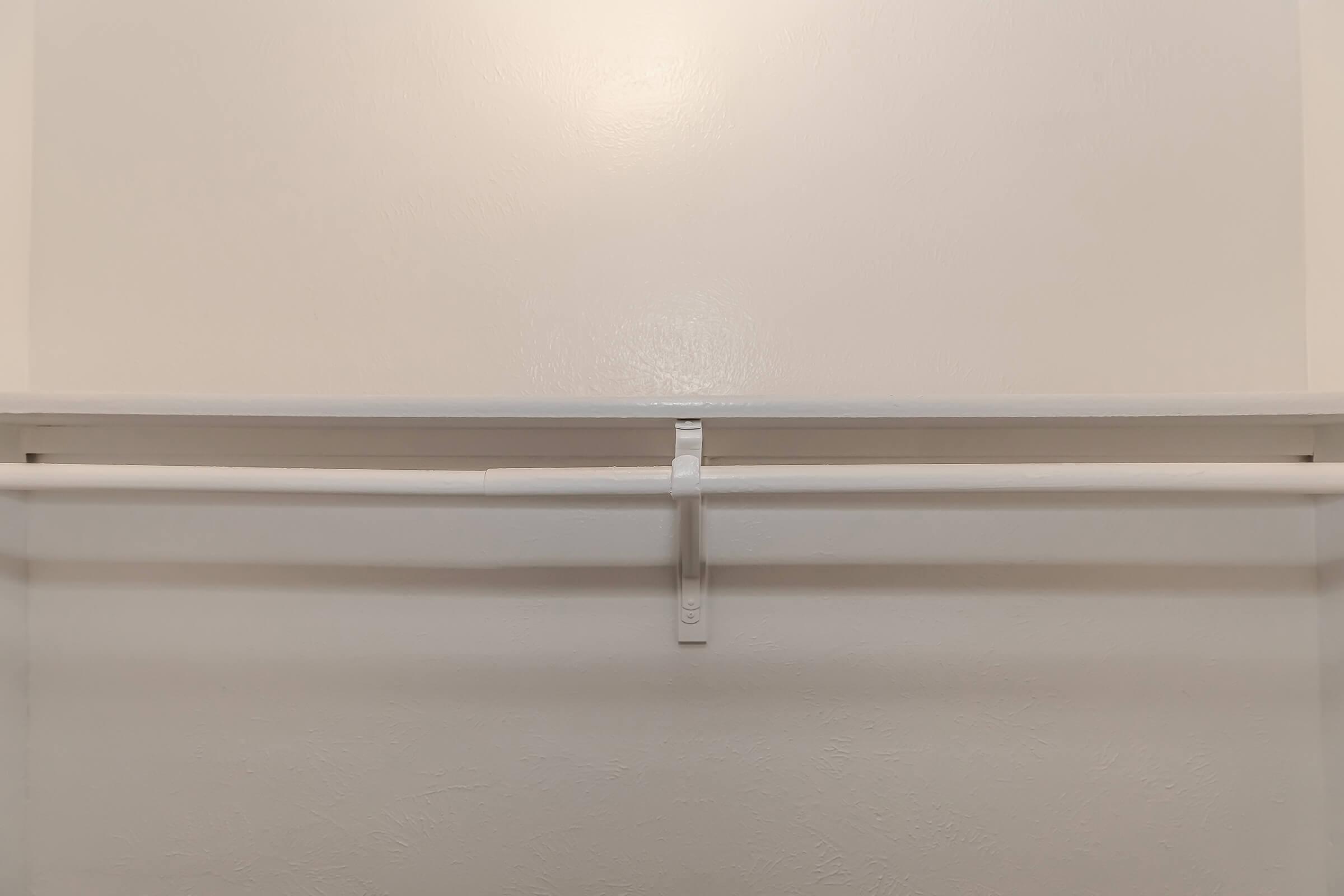 A view of an empty closet with a plain white wall and a horizontal white rod for hanging clothes. The rod is supported by a simple plastic bracket in the center, and the overall appearance is minimalistic and clean.