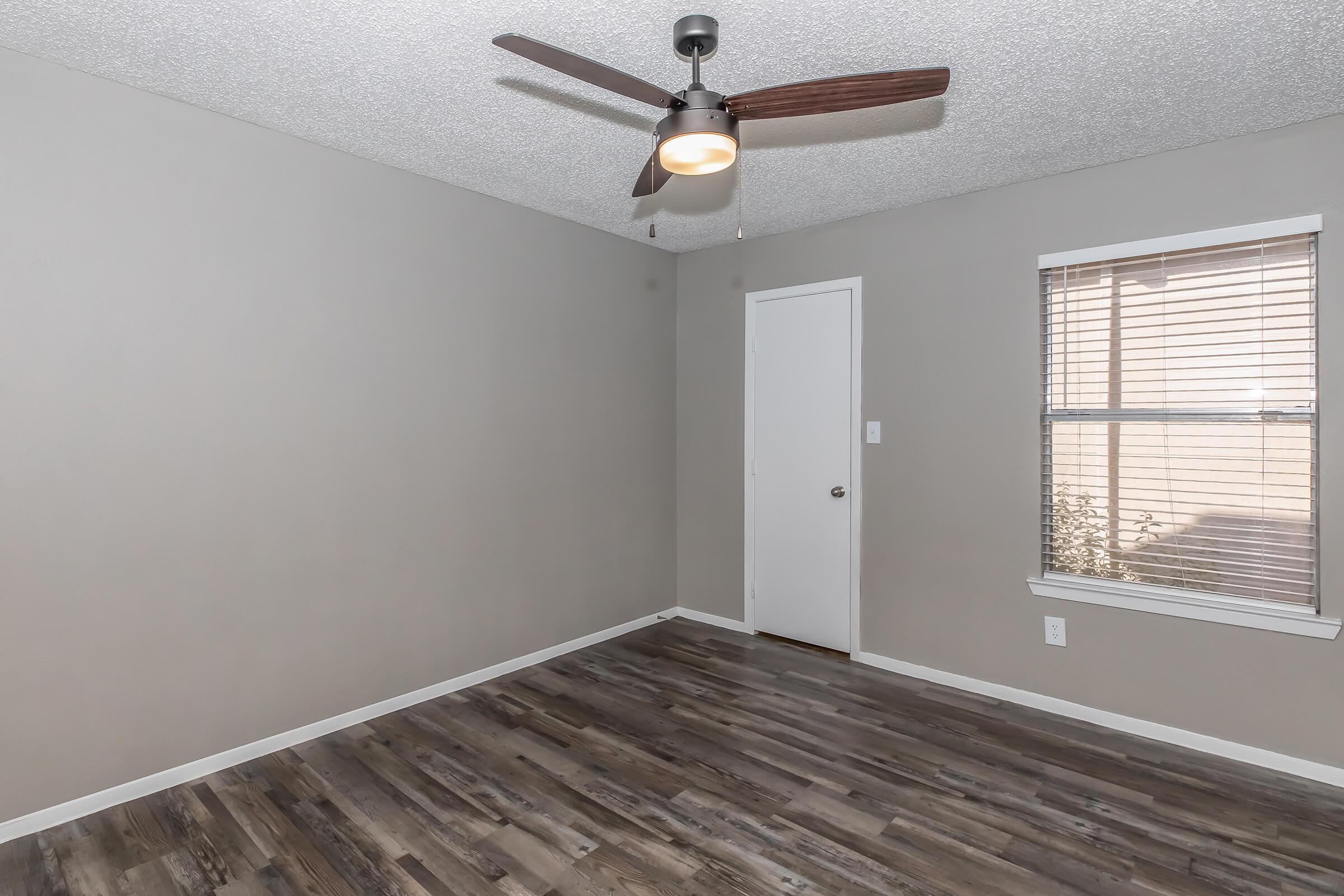 An empty room featuring light gray walls, a ceiling fan with wooden blades, and a window with blinds. The floor is covered in dark wooden laminate. A closed door is visible on one wall, and the room has a clean, minimalist aesthetic. Soft natural light filters through the window.