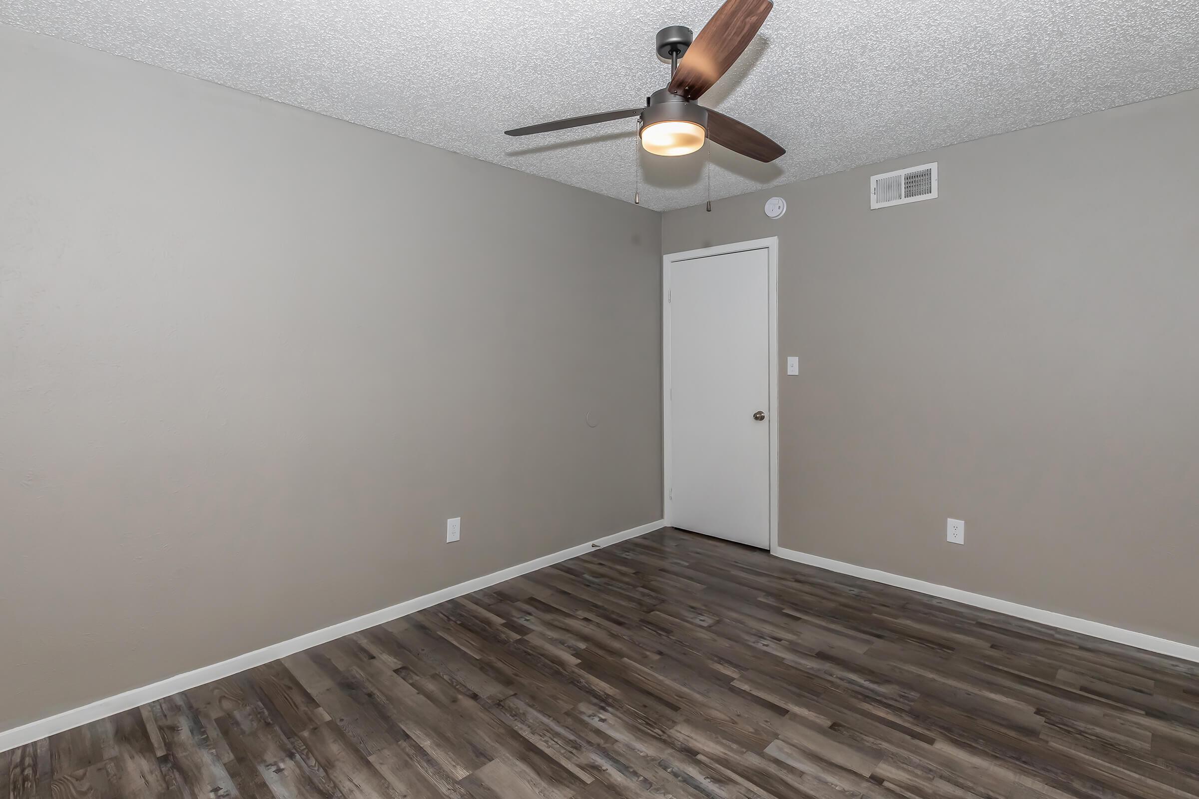 A sparsely furnished room with light gray walls and a textured ceiling. The floor features brown wood-like laminate. A ceiling fan with a light fixture is installed. A closed white door is visible, along with two light switches on the wall. The overall atmosphere is neutral and minimalist.