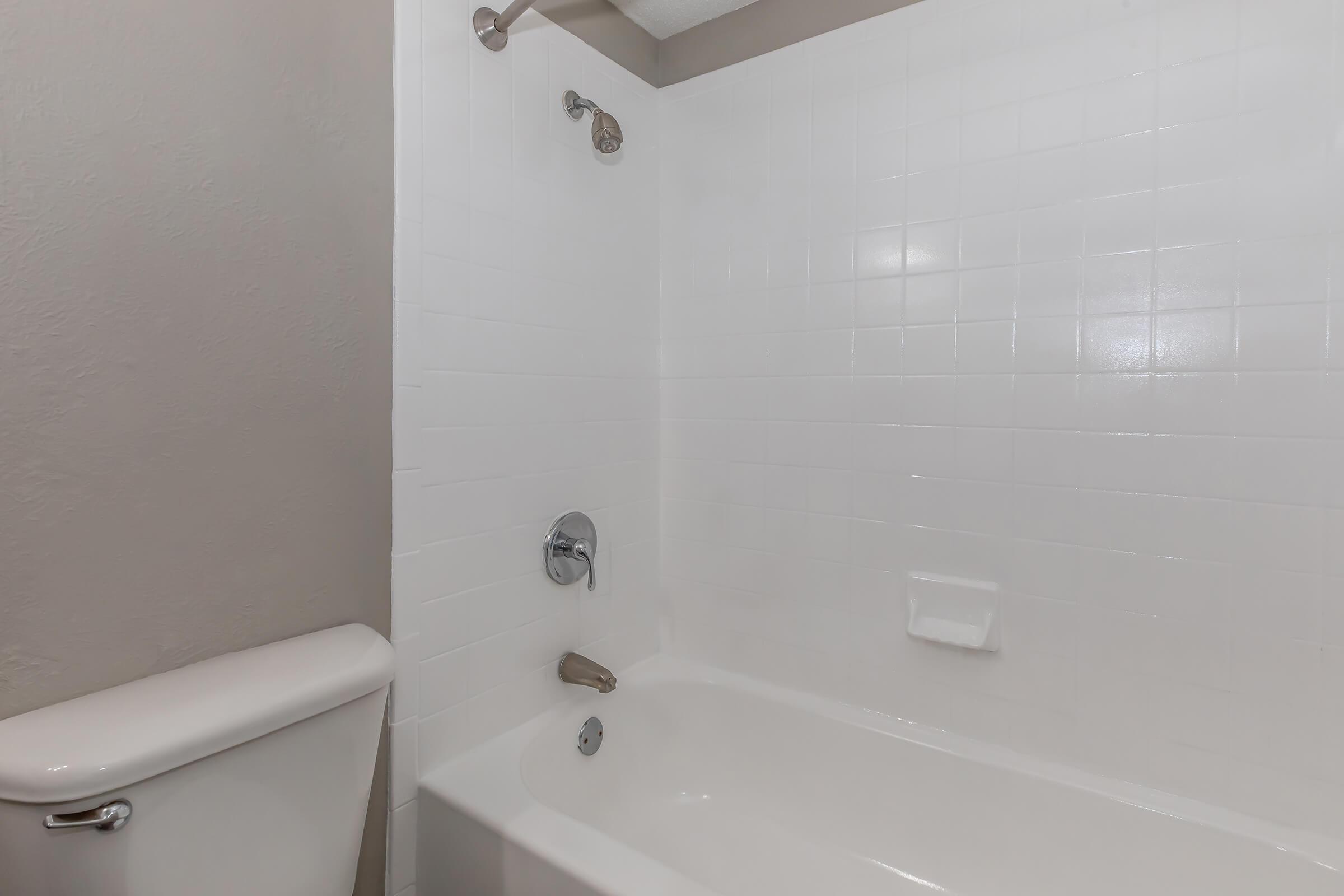 A clean and modern bathroom featuring a white tub with a chrome faucet, a wall-mounted showerhead, and white tiled walls. The scene includes a toilet to the left, with a neutral-colored wall in the background. The overall atmosphere is bright and minimalistic.