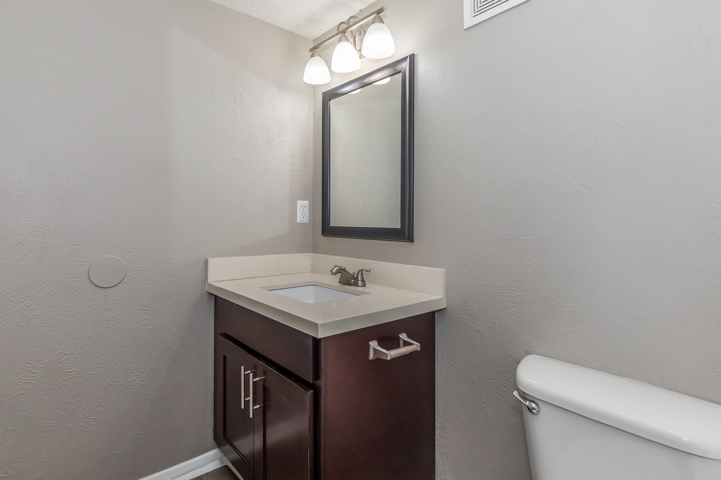 A small bathroom featuring a dark wood vanity with a sink, a rectangular mirror above it, a light fixture with three bulbs, a beige countertop, and a white toilet on the right against a textured gray wall. The space is well-lit and organized.