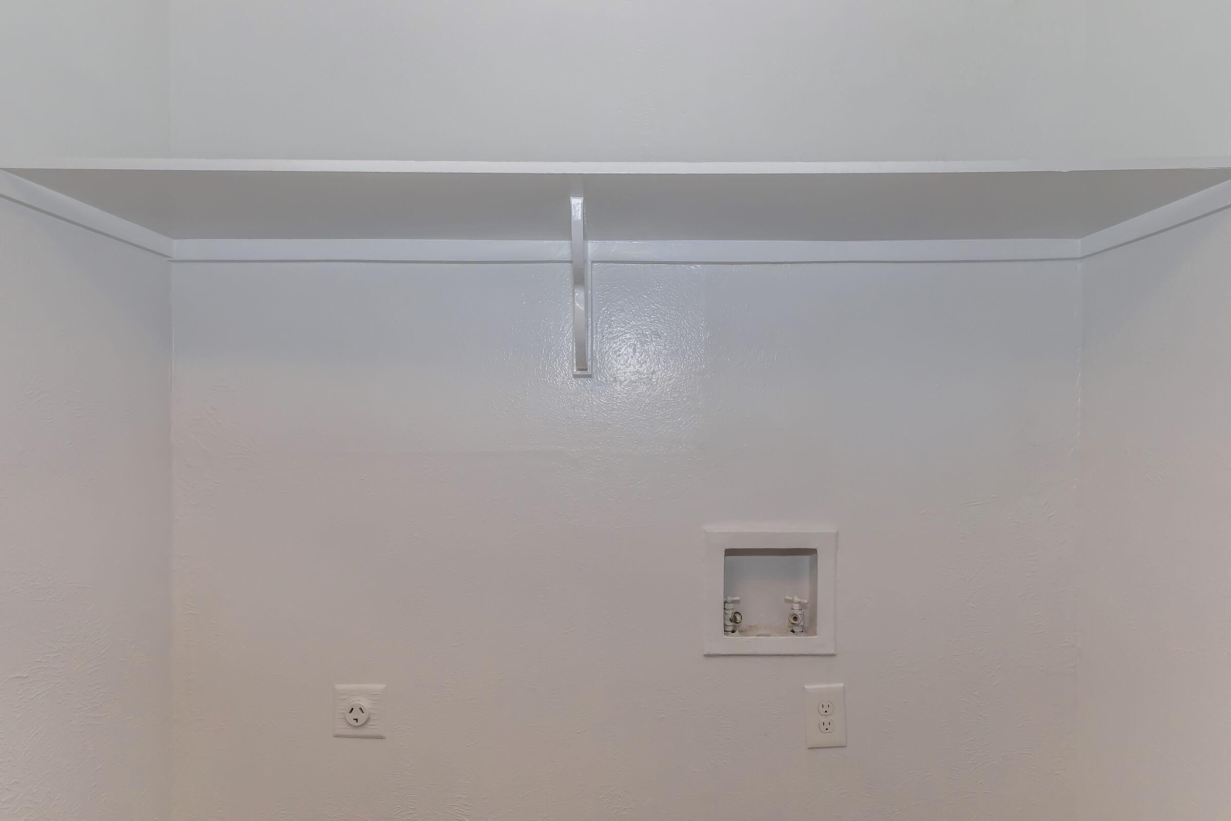 A simple, empty room with white walls and a shelf mounted on the wall. There are two electrical outlets visible on the wall, and a small recess that appears to hold fixtures or storage. The space is devoid of furniture or decorations, giving it a clean and minimalist appearance.