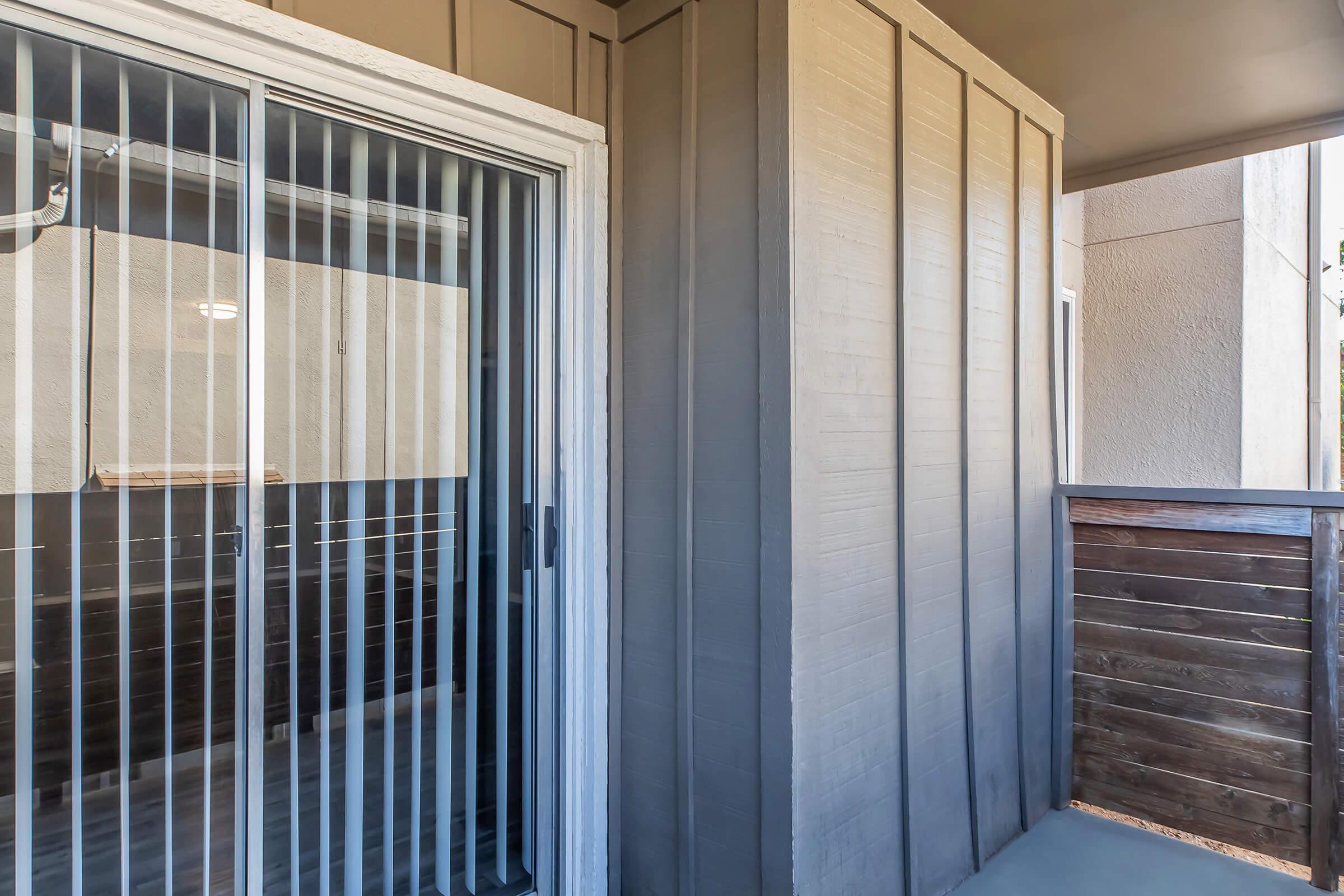 A balcony area featuring sliding glass doors with vertical blinds, gray walls, and wooden railing. The space is well-lit and has a modern design, suitable for outdoor relaxation or as an extension of interior living. The balcony offers a view of an adjacent area.