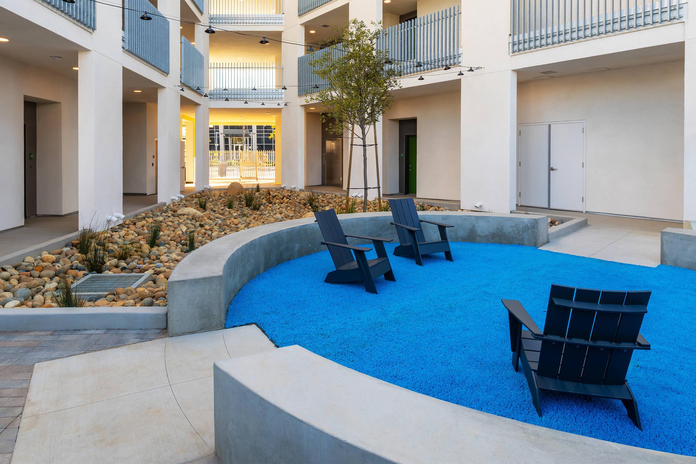 A courtyard featuring modern design with three dark blue lounge chairs arranged on bright blue gravel. Surrounding the seating area are smooth pebbles and small plants. The architecture consists of white walls and balconies, creating a clean and open atmosphere. Natural light brightens the space.