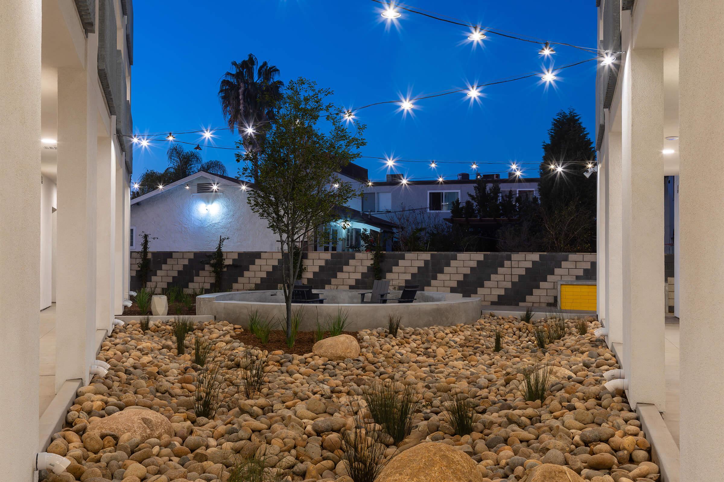 An outdoor courtyard with smooth river rocks scattered on the ground, surrounded by white walls. A tree stands in the center, and strings of lights crisscross above, glowing against a twilight sky. In the background, a house and garden are visible, adding depth to the serene atmosphere.