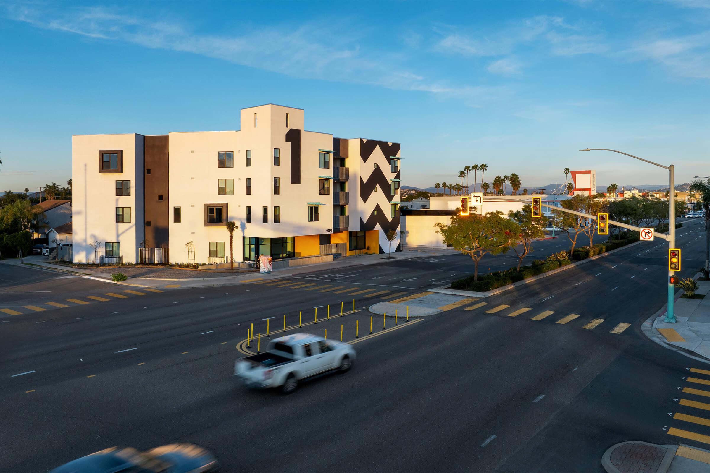 A modern building with a geometric black and white design, situated at a city intersection. The scene includes a clear blue sky, palm trees, and a white vehicle approaching the crosswalk. Yellow lane markings and traffic signals are visible, indicating an urban setting.