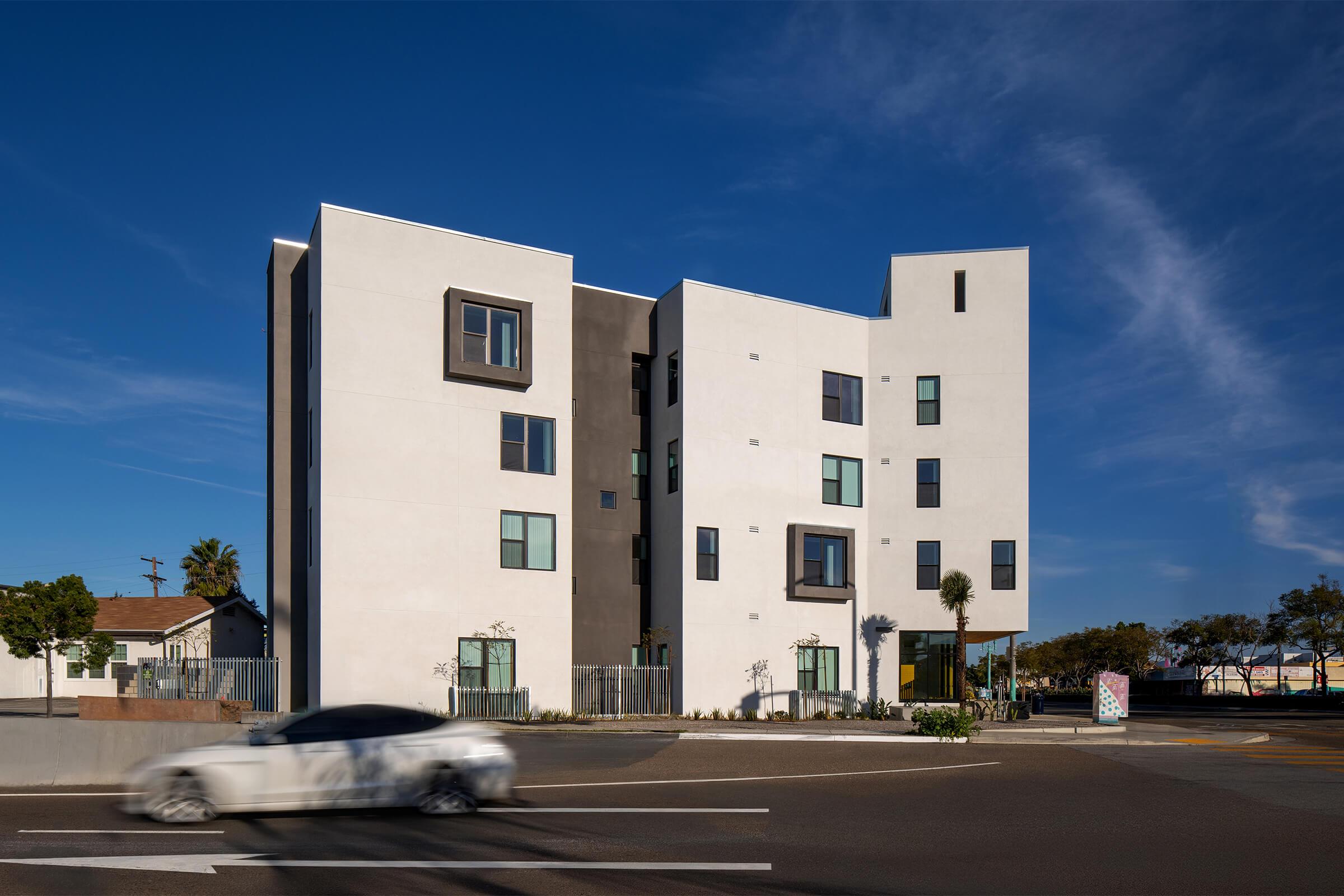 A modern, multi-story white apartment building with large windows and geometric design elements. The building is set against a clear blue sky, and a car is seen moving past it on the street. In the foreground, there are landscaped areas and minimal surrounding buildings.
