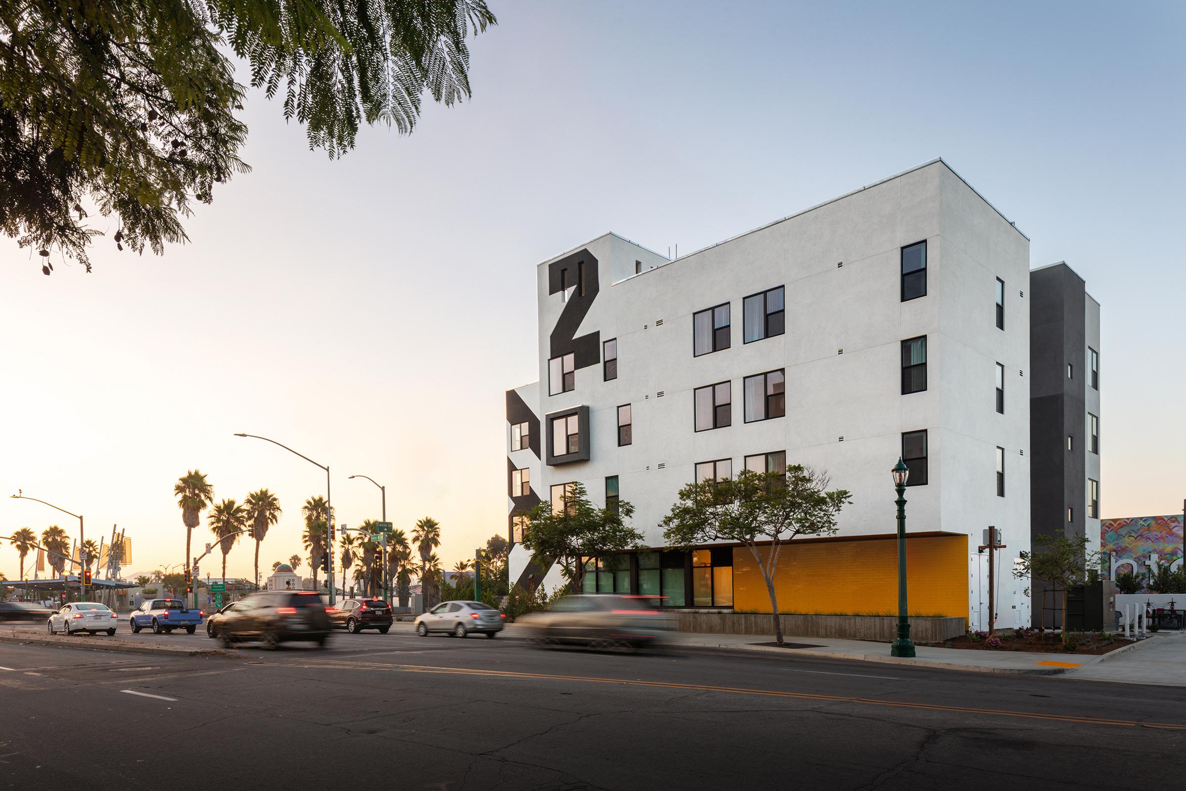 A modern multi-story building with a white exterior and large black number "2" prominently displayed. The lower section features bright orange accents, and palm trees line the street. Vehicles are passing by, and the scene is set during sunset, creating a vibrant urban atmosphere.