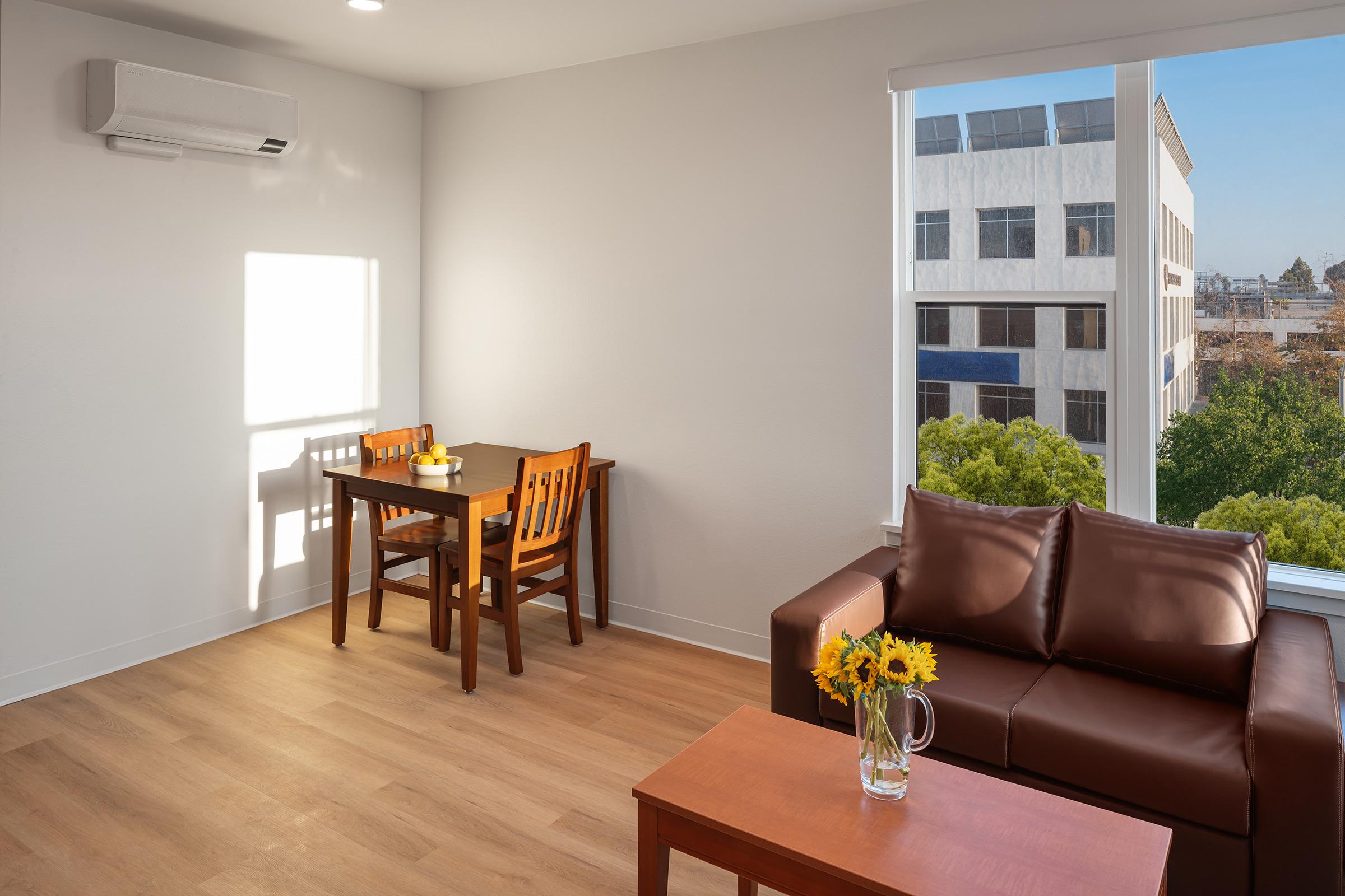 A cozy modern living space featuring a brown leather sofa, a wooden dining table with chairs, and a window with a view of an outdoor landscape. Sunlight streams in, illuminating the room, which also has a small vase of sunflowers on the coffee table.