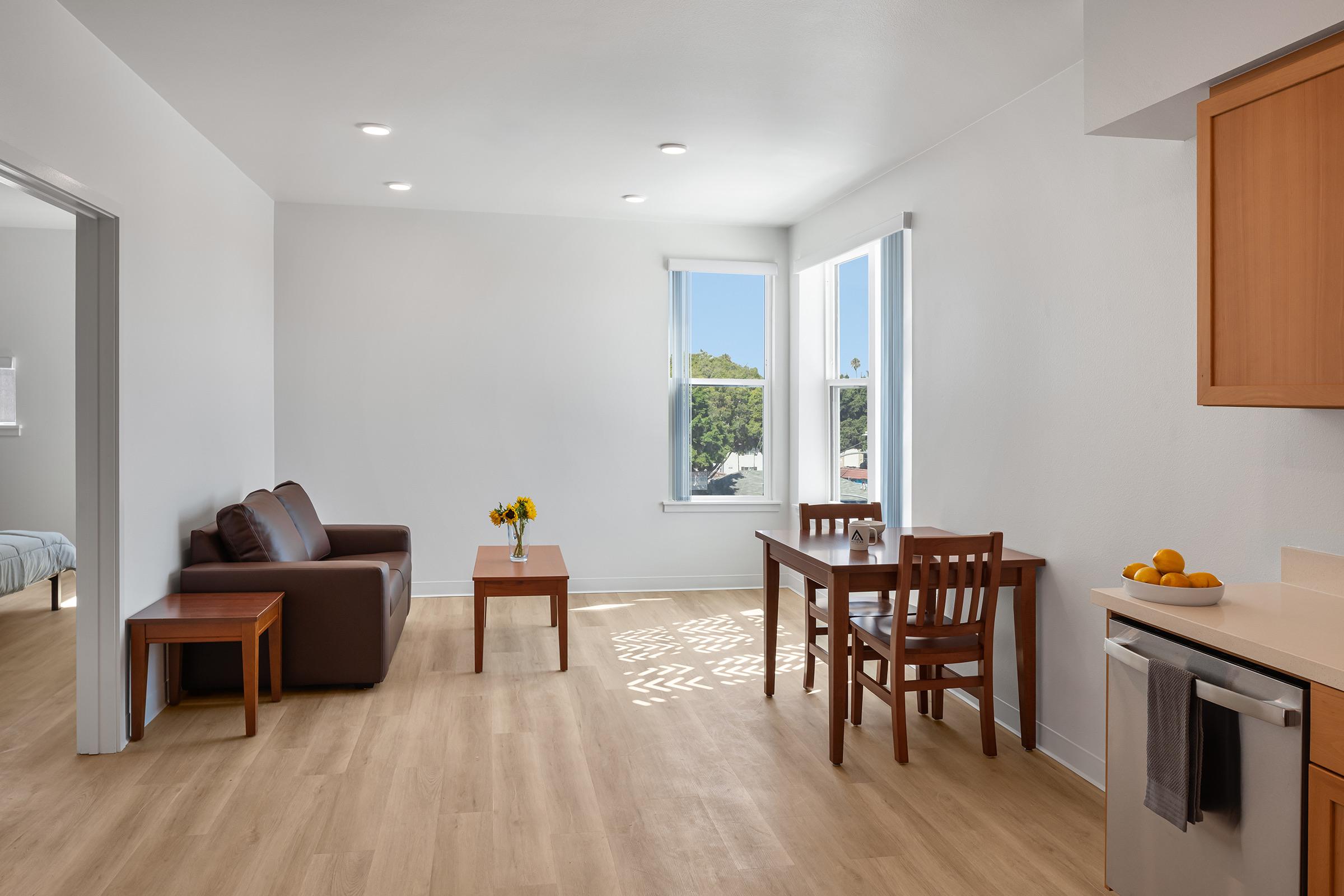 A modern, well-lit living space featuring a brown leather sofa, a wooden coffee table with a bouquet of yellow flowers, a dining table with chairs, and a kitchen area. Large windows allow natural light to fill the room, highlighting the light wooden flooring and minimalist decor.