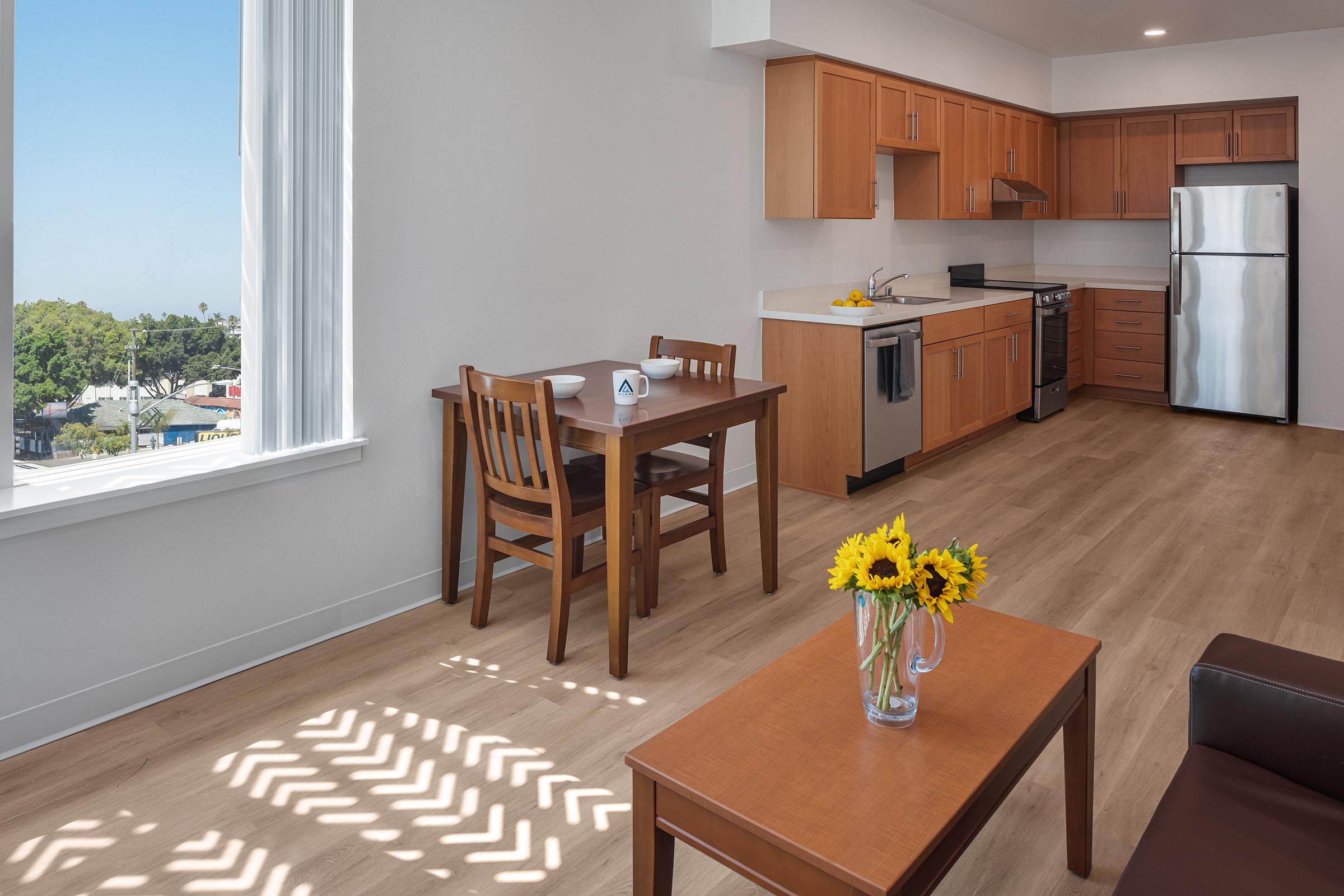 A bright and modern kitchen and dining area featuring wooden cabinets, stainless steel appliances, a small dining table with two chairs, a coffee table with a vase of sunflowers, and large windows that let in natural light, offering a view of greenery outside.