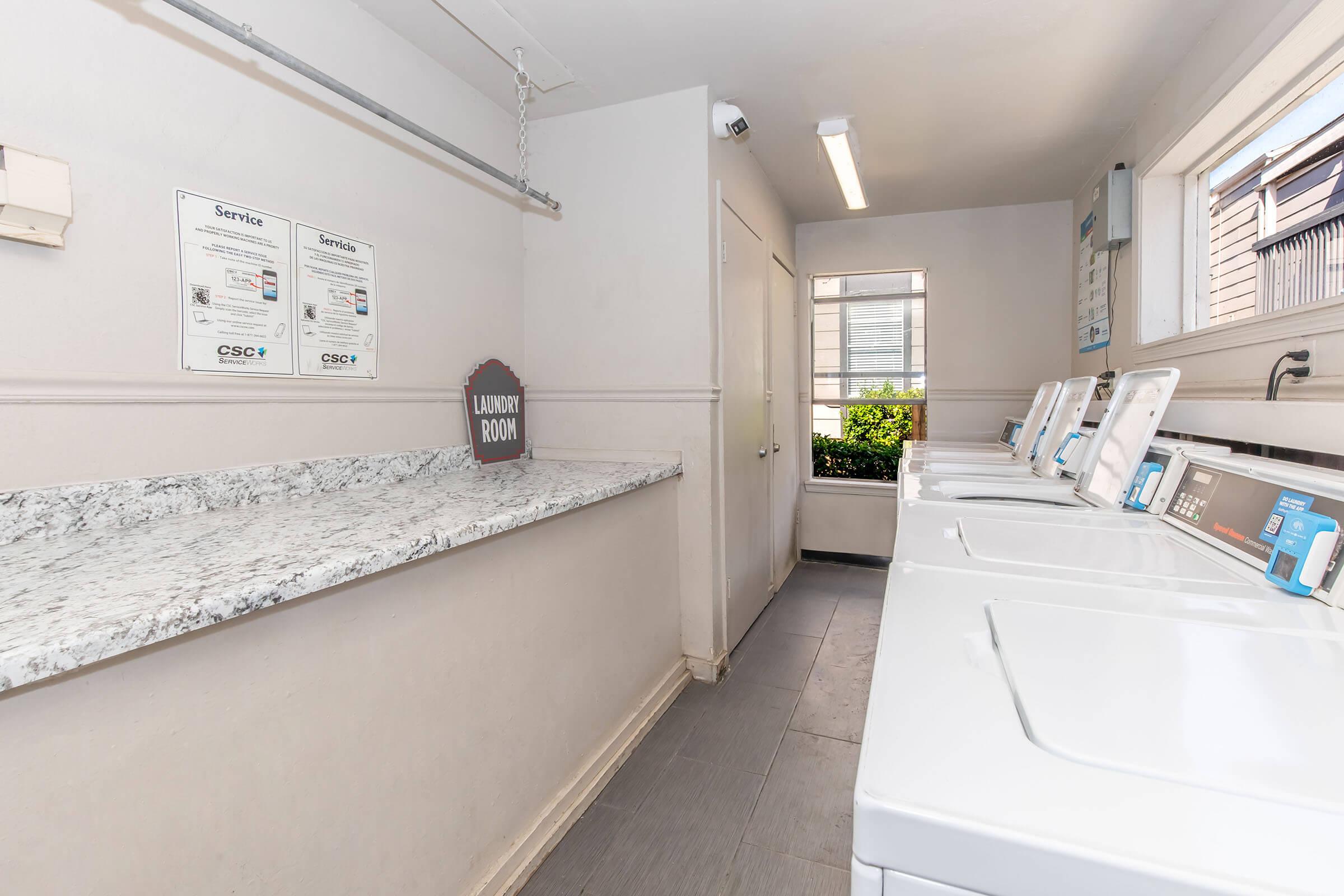 A clean and bright laundry room featuring several white washing machines and a countertop made of granite. Service signs are displayed on the wall, and a door leads outside to a windowed area with greenery. The space is well-lit with a modern and tidy appearance.