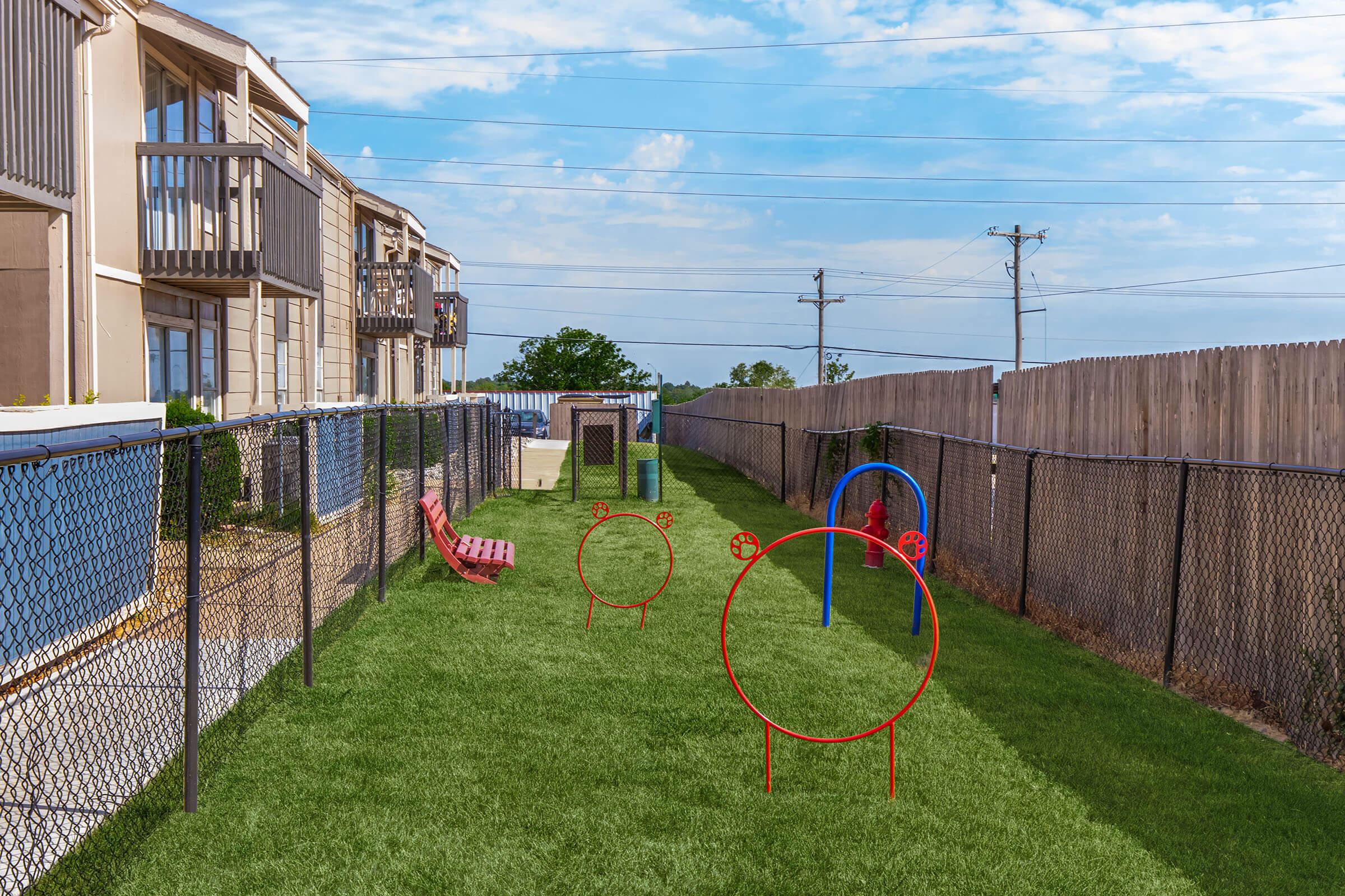 A grassy backyard area featuring a fenced space. Colorful circular play structures in red and blue are positioned on the grass, with some lounge chairs nearby. The backdrop includes two multi-story buildings and a wooden fence, under a partly cloudy sky. Power lines can be seen in the distance.