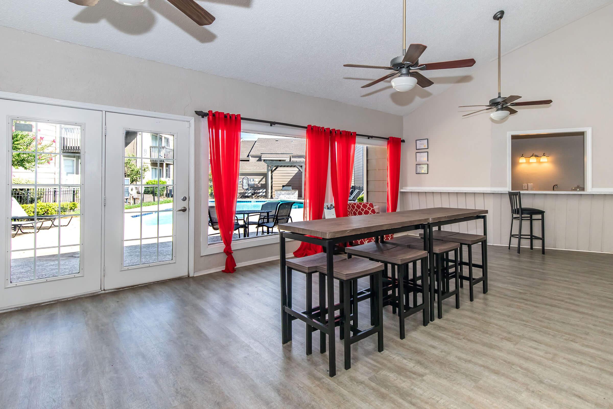 A bright, spacious room with large windows featuring red curtains, showcasing a view of an outdoor pool area. There is a long dining table with high stools, two ceiling fans, and adjacent to a bar area with seating. The walls are neutral-colored, creating a modern, inviting atmosphere.